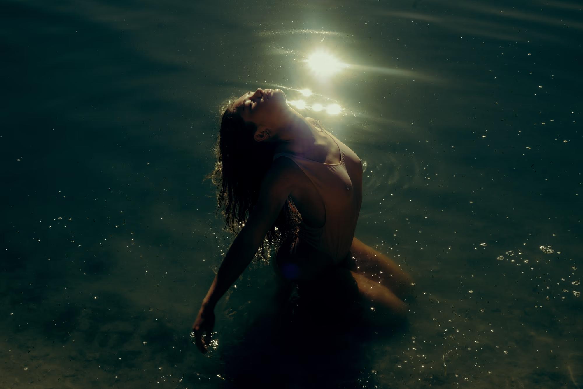 Underwater wide shot of woman swimming beneath lake surface with sunlight halo effect, dark teal tones, cinematic fine art