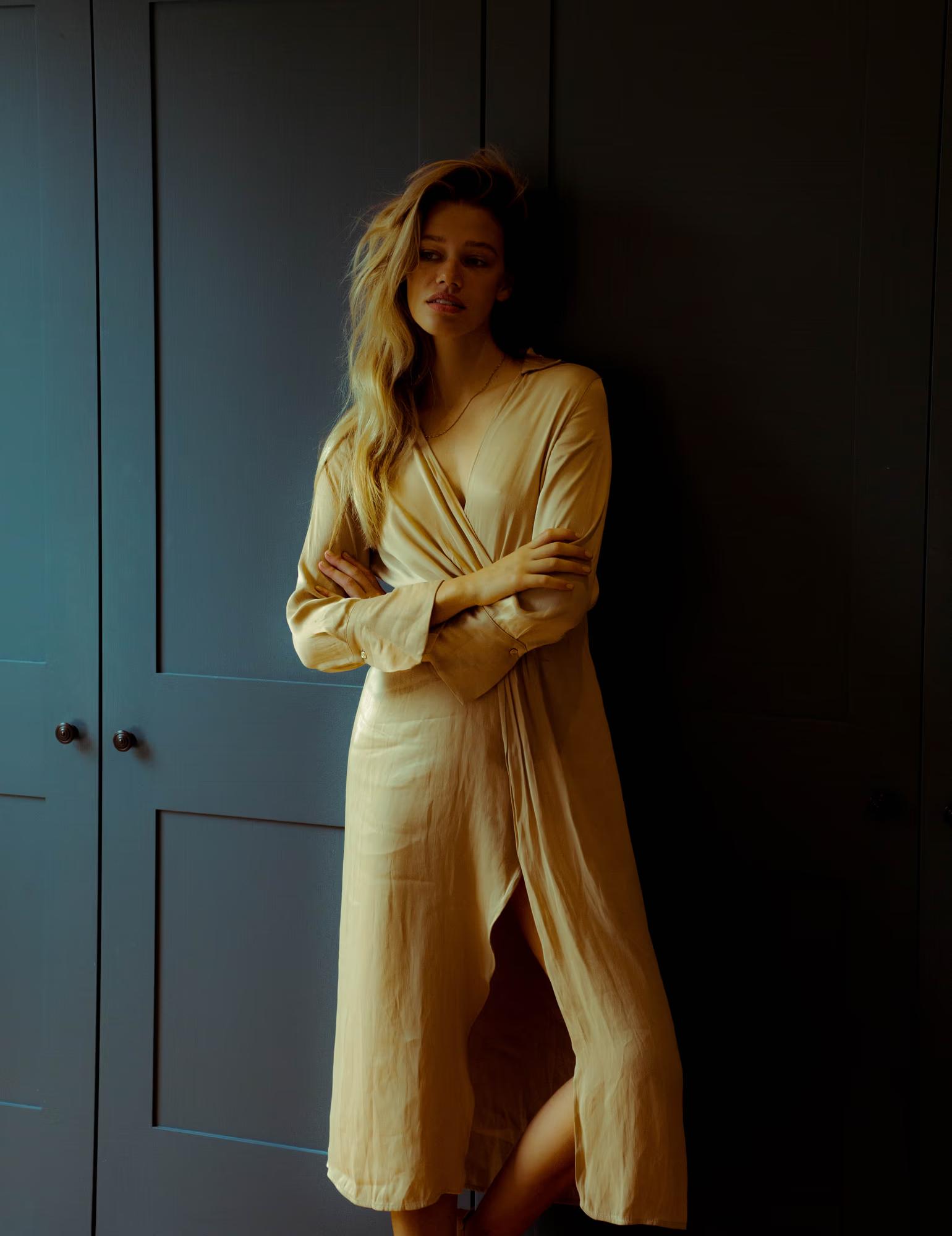A model with long wavy blonde hair leans against a dark teal wardrobe, arms crossed, looking downward. She wears a flowing sand-coloured wrap midi dress with long sleeves. Warm directional light catches her hair and the silky fabric against the deep shadow behind her.