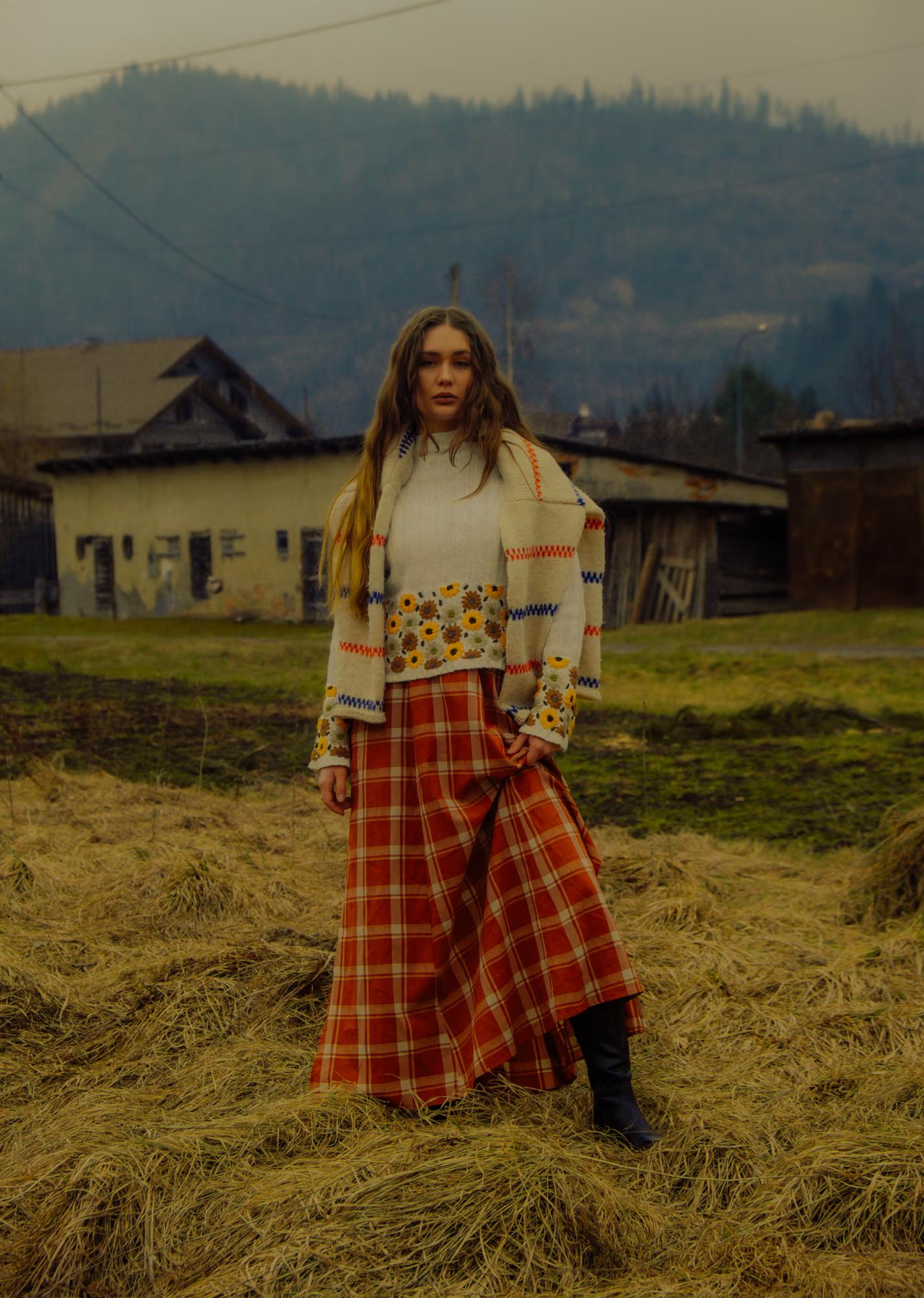 Full body fashion editorial shot of woman in white blouse and red tartan maxi skirt standing in Carpathian mountain meadow with traditional wooden house in background, Ukrainian ethno style