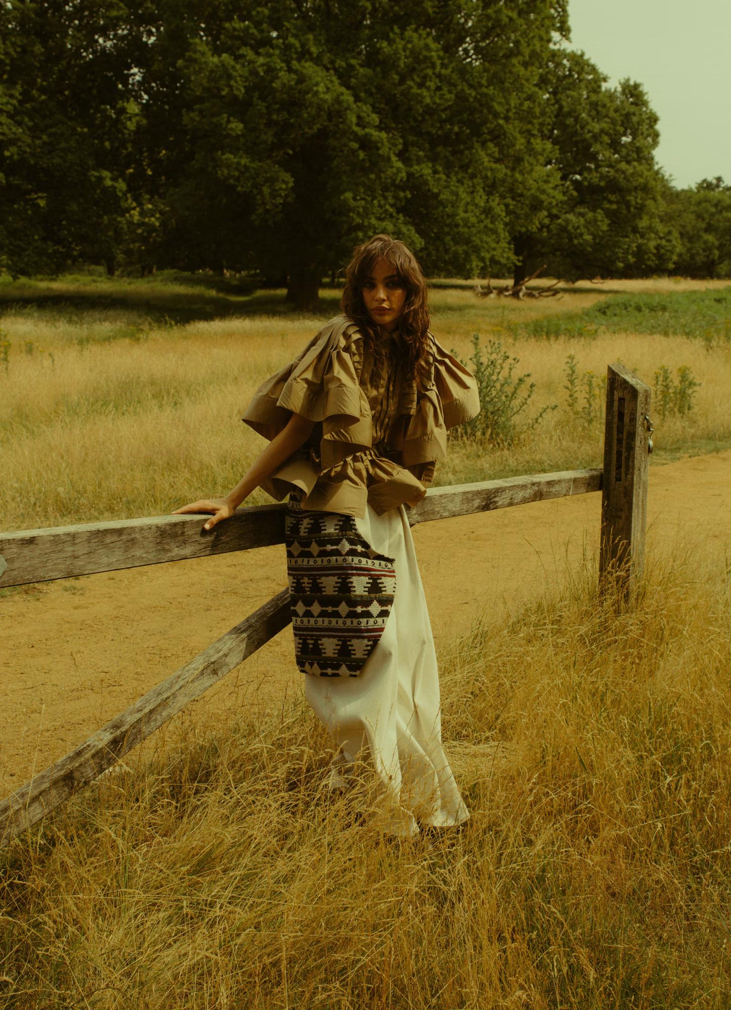 A model leans against a weathered wooden fence post on a dusty path at the edge of a golden field. She wears a voluminous tan ruffled-shoulder top and a woven geometric-patterned midi skirt in navy, white, and red. Her wavy dark hair falls loosely and she gazes at the camera.