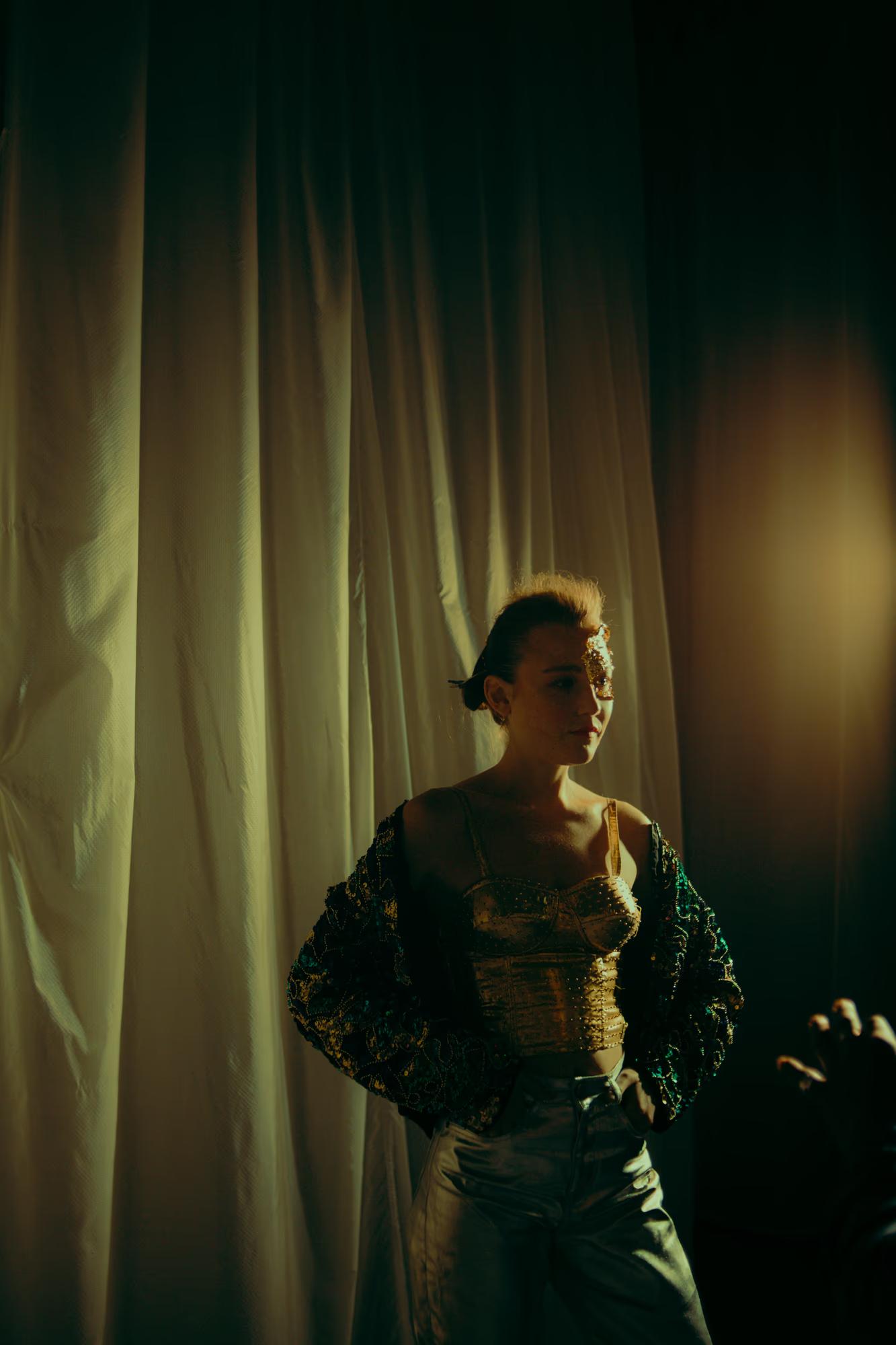 A model posing in a structured designer outfit under a singular spotlight in a dark backstage corridor.