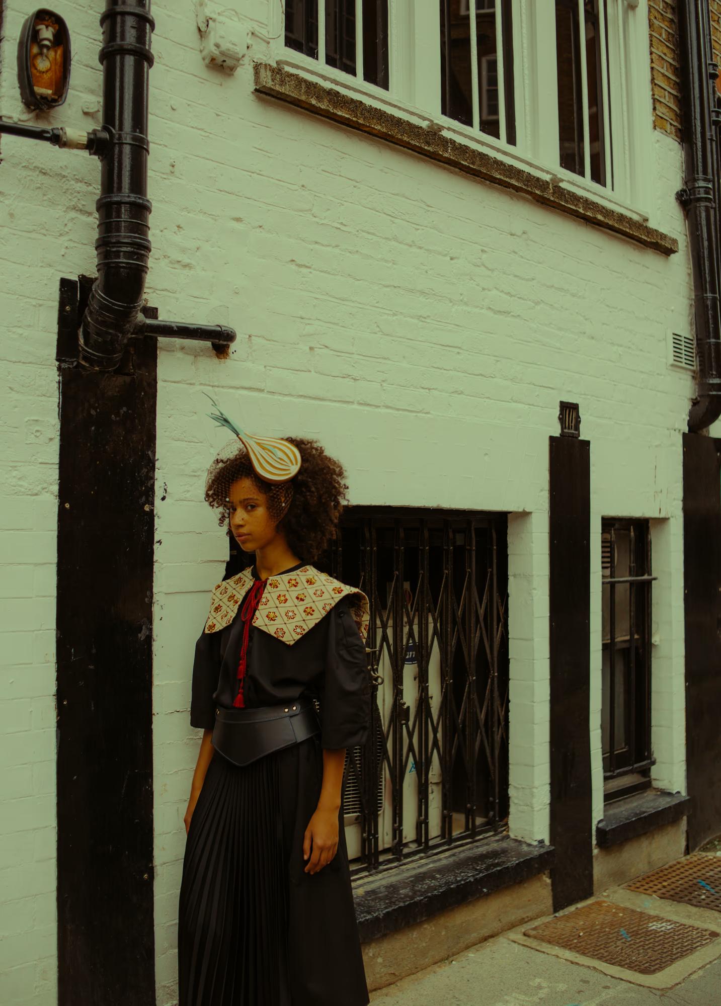 A model stands beside a black drainpipe against a white-painted brick building on a London street. She wears a black jacket, an embroidered wide collar with floral motifs, a red tie, a structured leather belt, and a long black pleated skirt. Perched on her natural curly hair is a quirky striped teardrop-shaped fascinator with a feather.