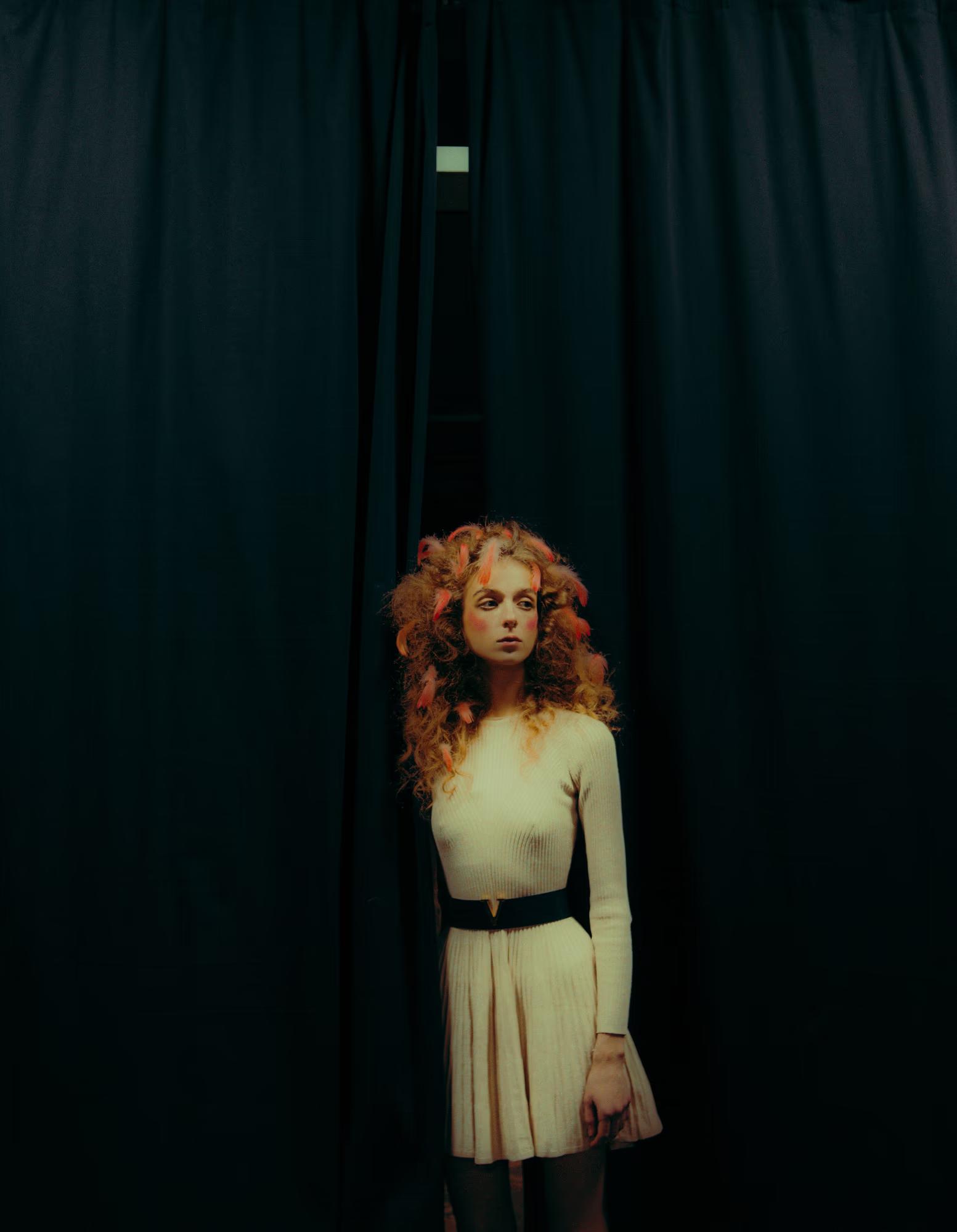 Full-length editorial photo of a woman in a pale gown standing in dramatic theater shadows.