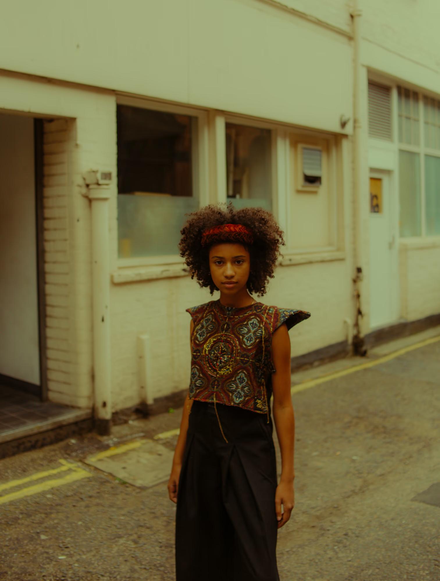 Close-up street portrait of a model with a full natural afro and a red headband, wearing a richly embroidered multicolour crop top with geometric and medallion motifs and a black wrap skirt. She stands in a quiet London backstreet, facing the camera with a composed expression.
