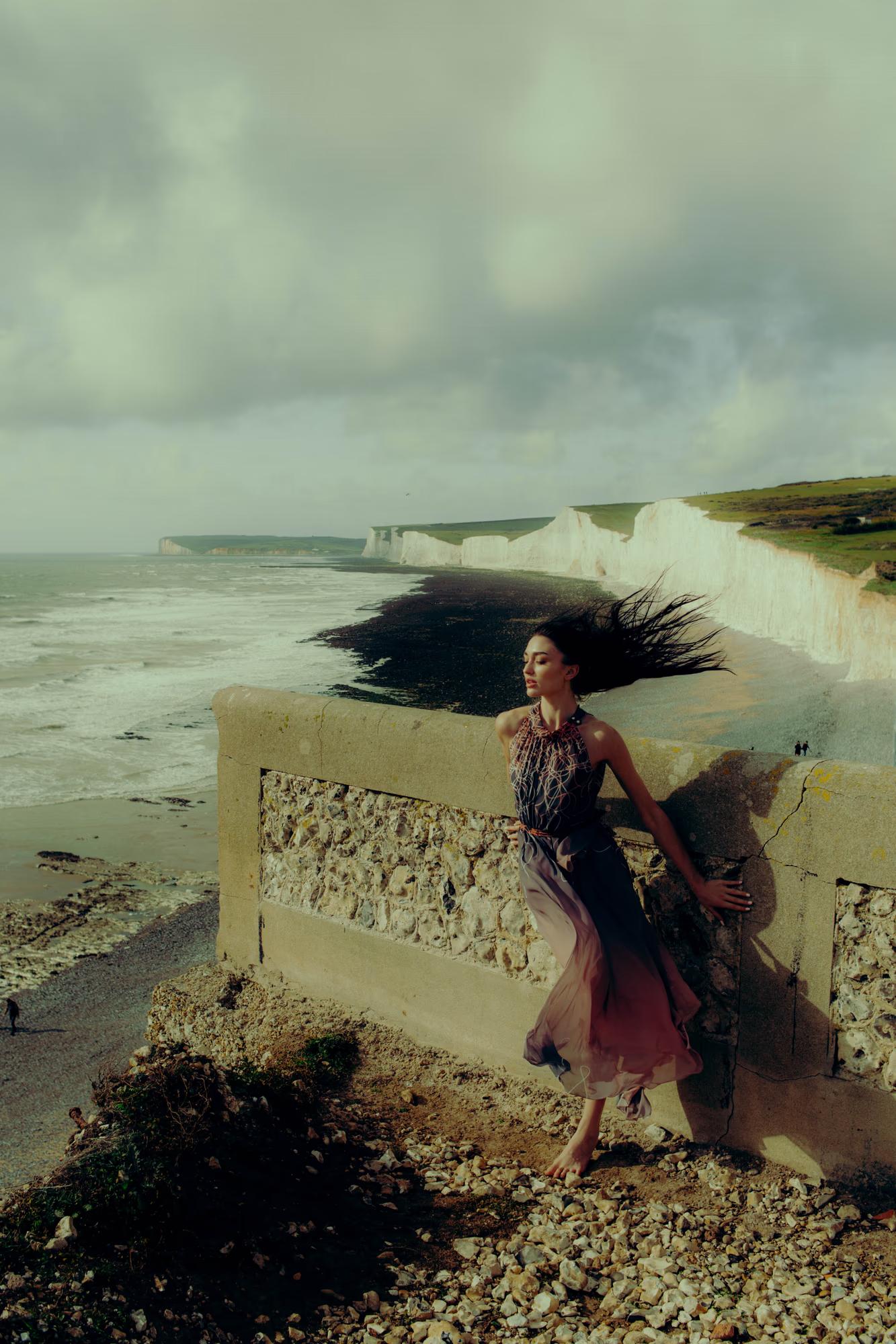 Full body fashion campaign shot of woman in dark designer outfit on Eastbourne rocky shore with dramatic stormy sky, moody coastal editorial