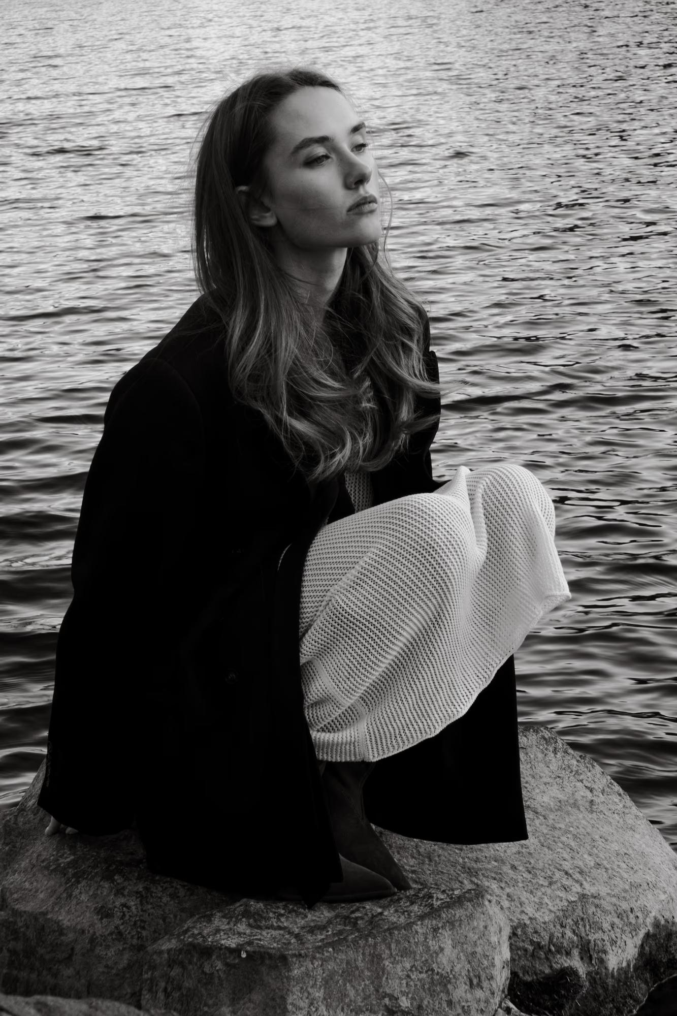 Dynamic black and white photograph of the model looking out over the expansive lake at sunset, high contrast.