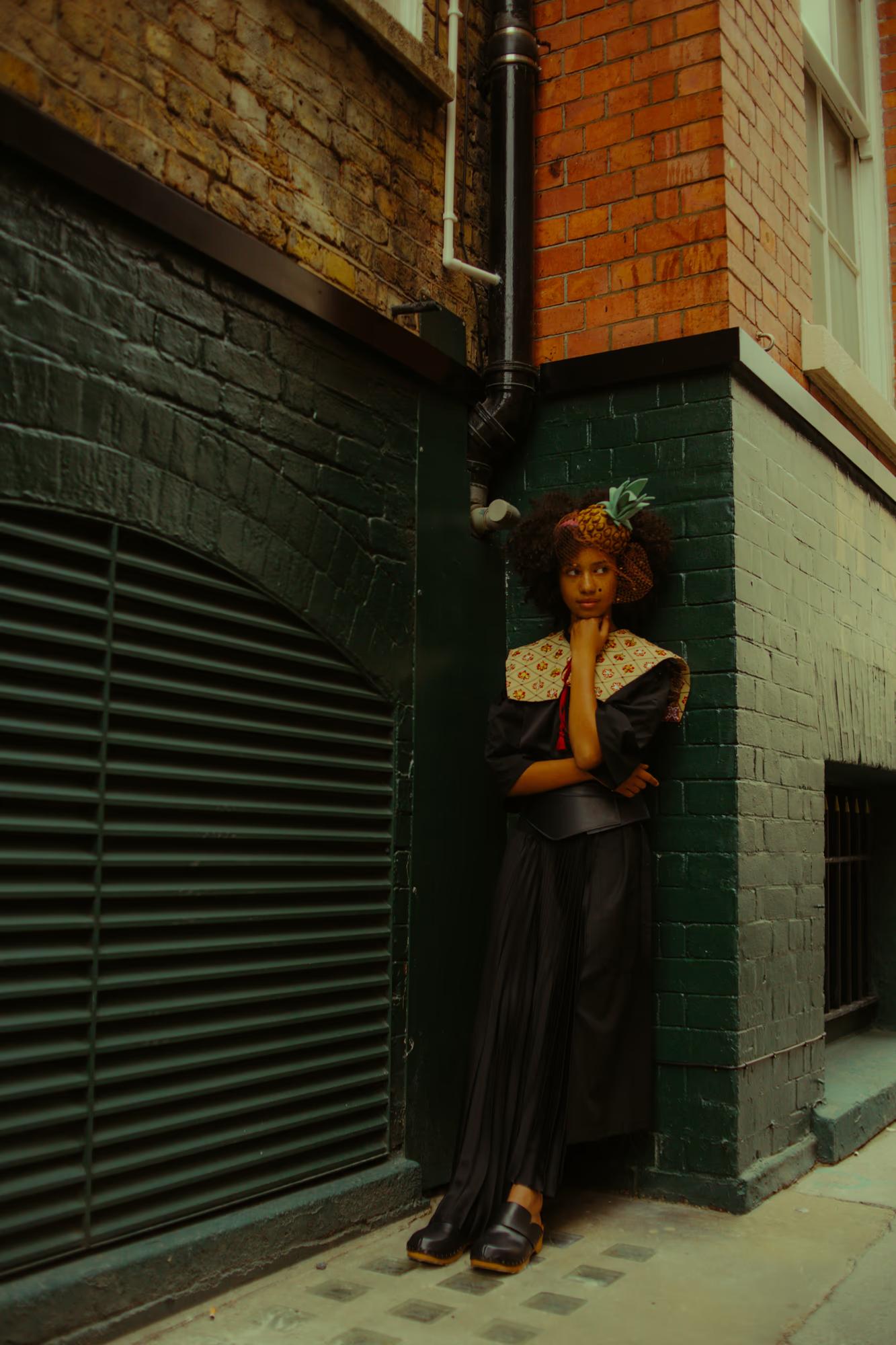 A model leans against the corner of a dark green-painted brick building in a London alley, one hand raised to her chin in a thoughtful pose. She wears a black outfit with an embroidered folk collar, a leather belt, a black pleated skirt, and chunky wooden clogs. On her head sits a small decorative headpiece resembling a miniature pineapple with leaves.