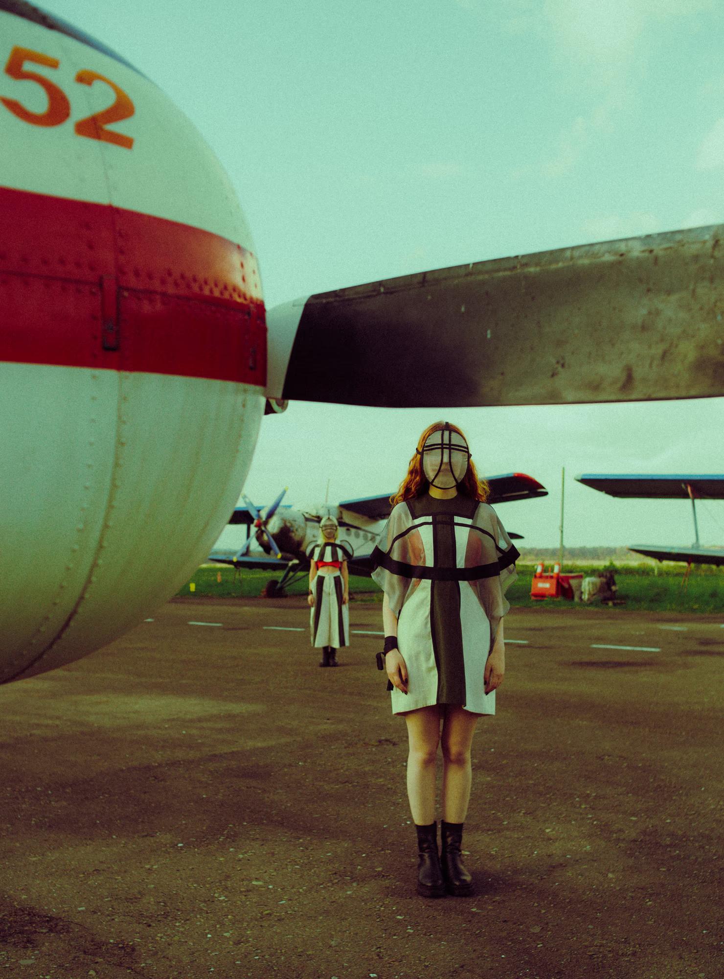 A model wearing a geometric black-and-white mini dress and a sheer grid-patterned face mask walks across an airfield tarmac. The large nose section of a plane marked "52" dominates the left side of the frame. A second similarly dressed figure is visible in the distance behind her.