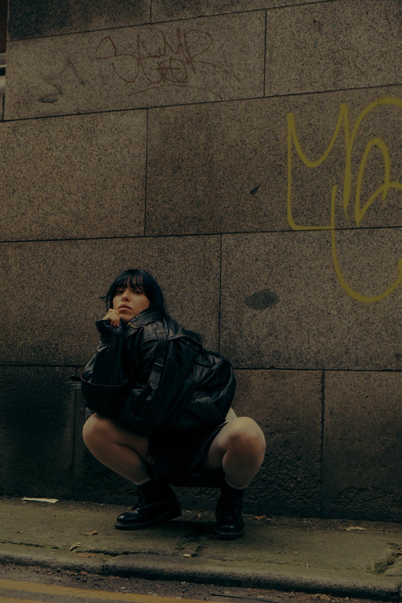 Full body fashion shot of person in dark outfit crouching on Manchester street pavement, warm sepia tones, gritty urban editorial