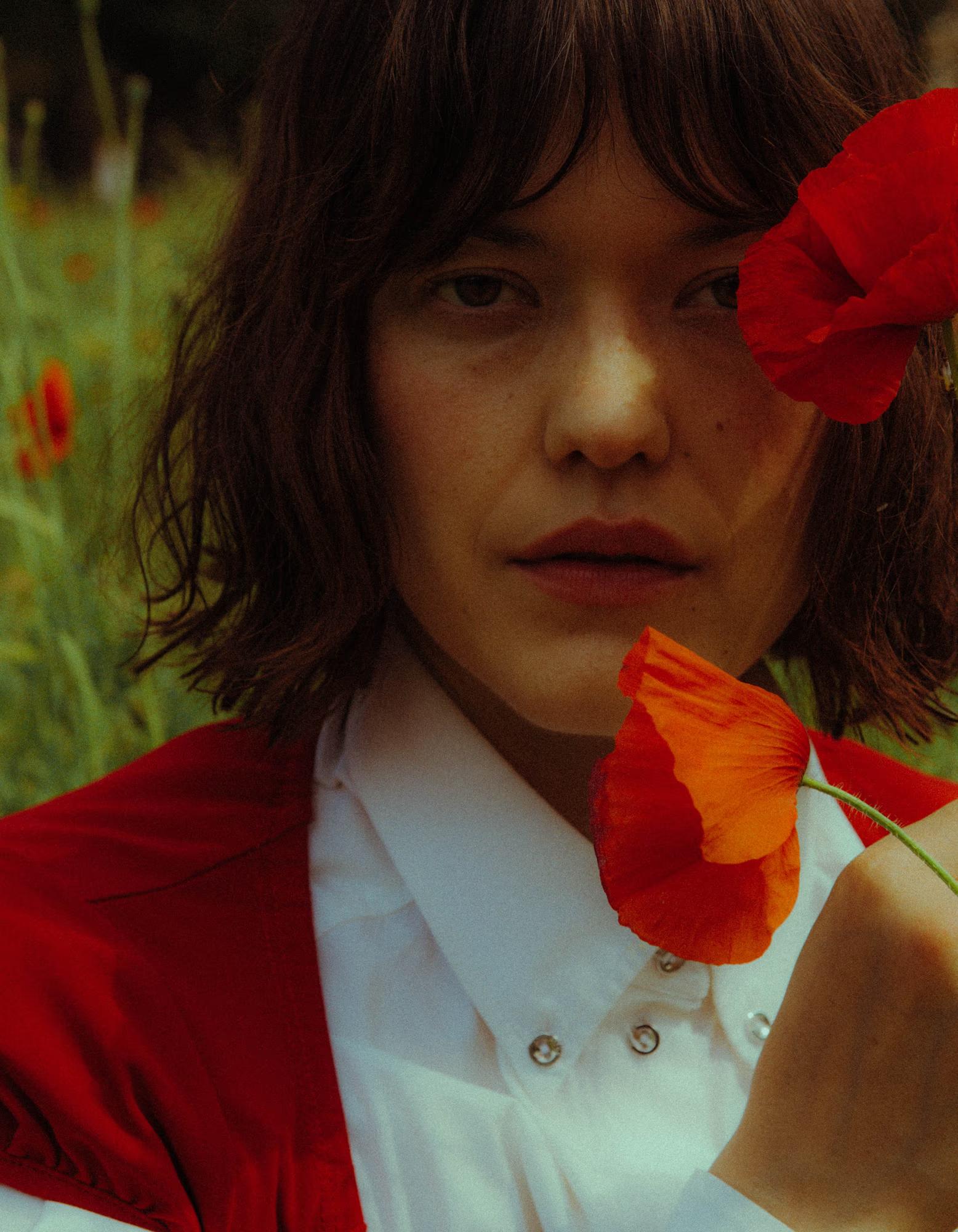 Close-up portrait of a model with a dark bob and fringe, holding two red poppies — one beside her eye, one at her chin — and gazing directly into the camera. She wears a white collared shirt layered under a vivid red garment. A blurred poppy field is visible in the background.