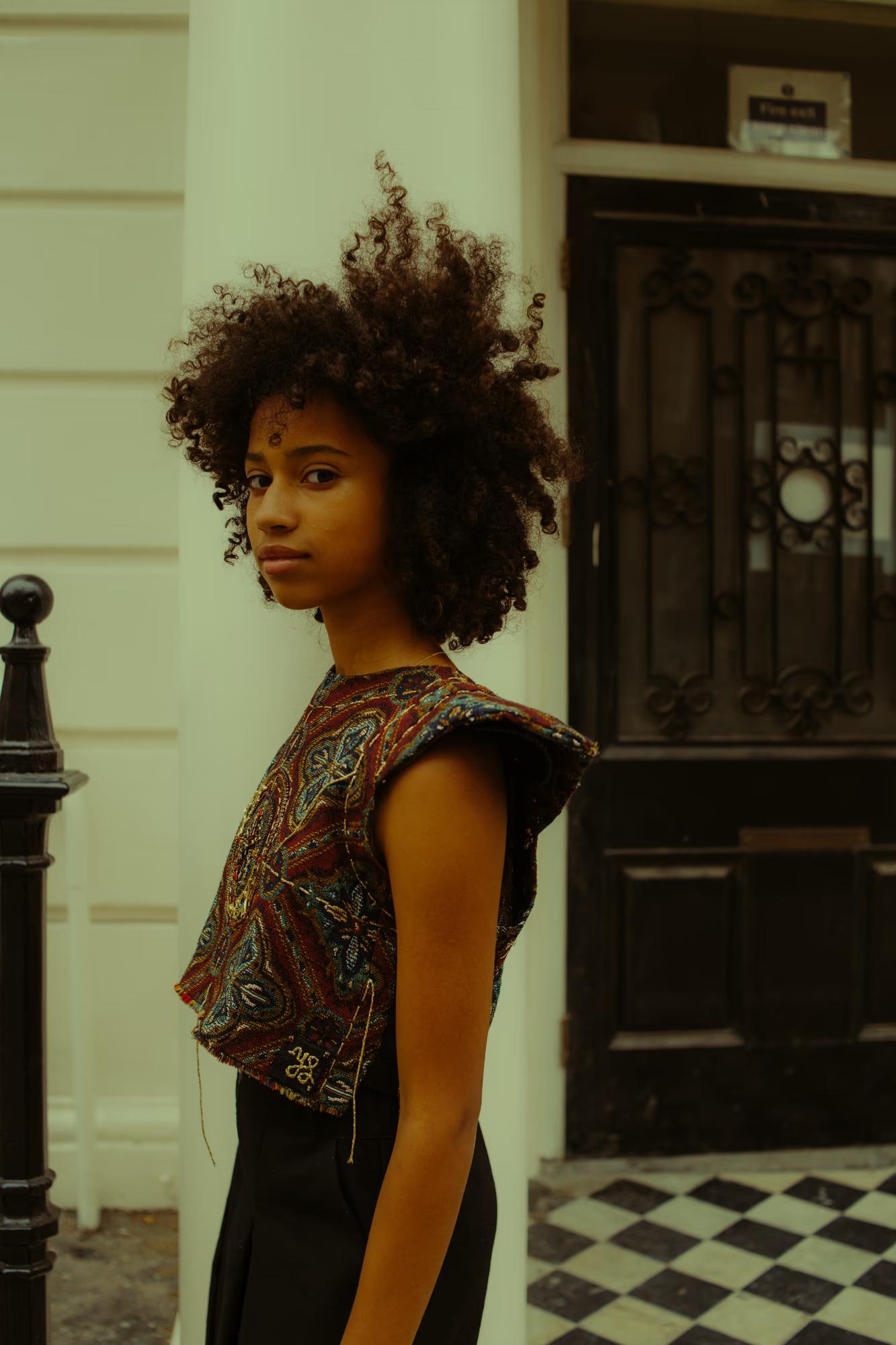 Side-profile close-up of a model with a voluminous natural afro, wearing a heavily embroidered short-sleeve top in jewel tones — deep red, teal, and gold — over a black skirt. She glances back over her shoulder toward the camera. A black iron door with ornate detailing and a black-and-white tiled step are visible behind her.
