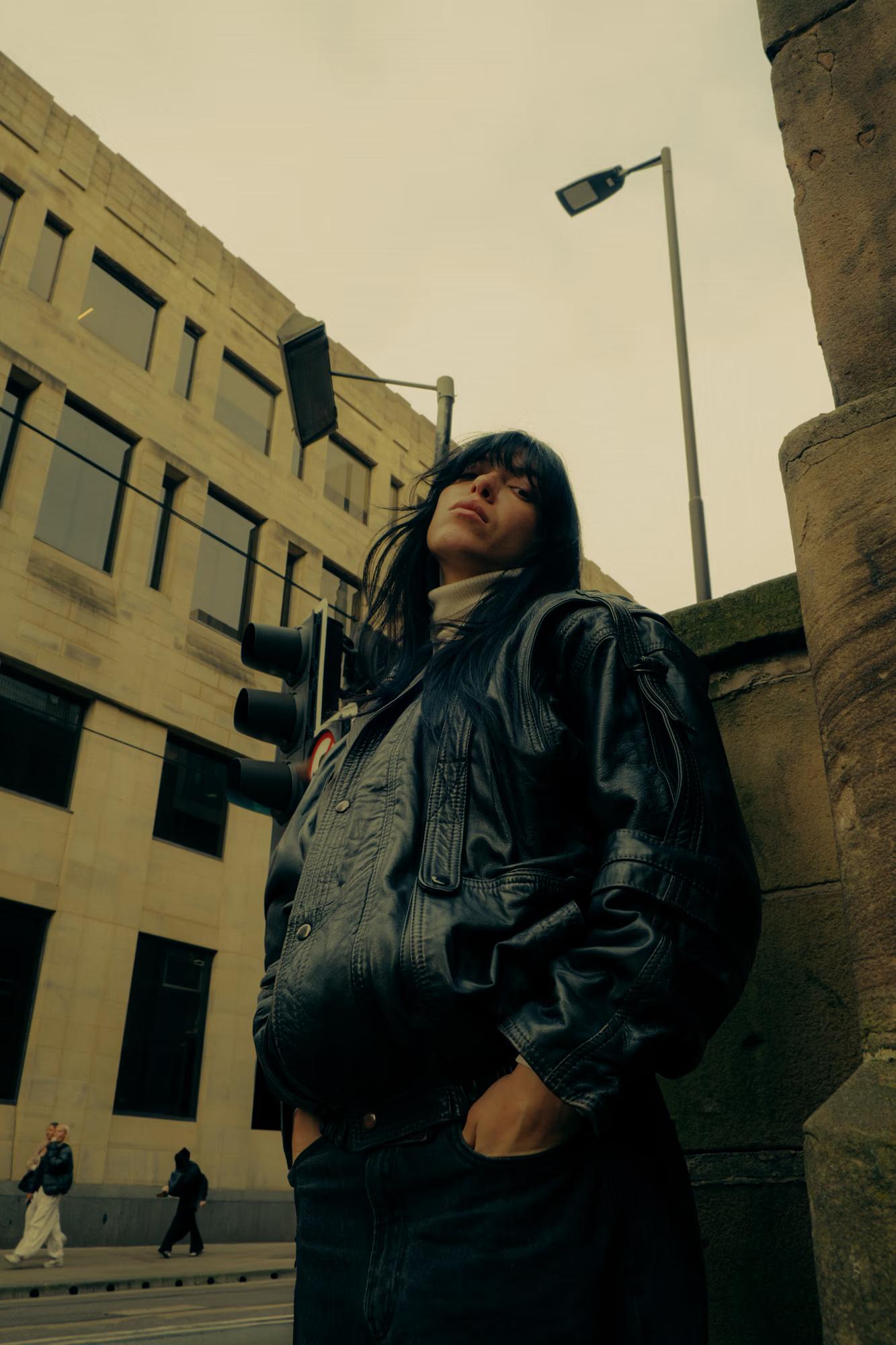 Full body fashion shot of person in dark oversized coat and cap walking on Manchester street with brutalist buildings in background, sepia toned urban editorial