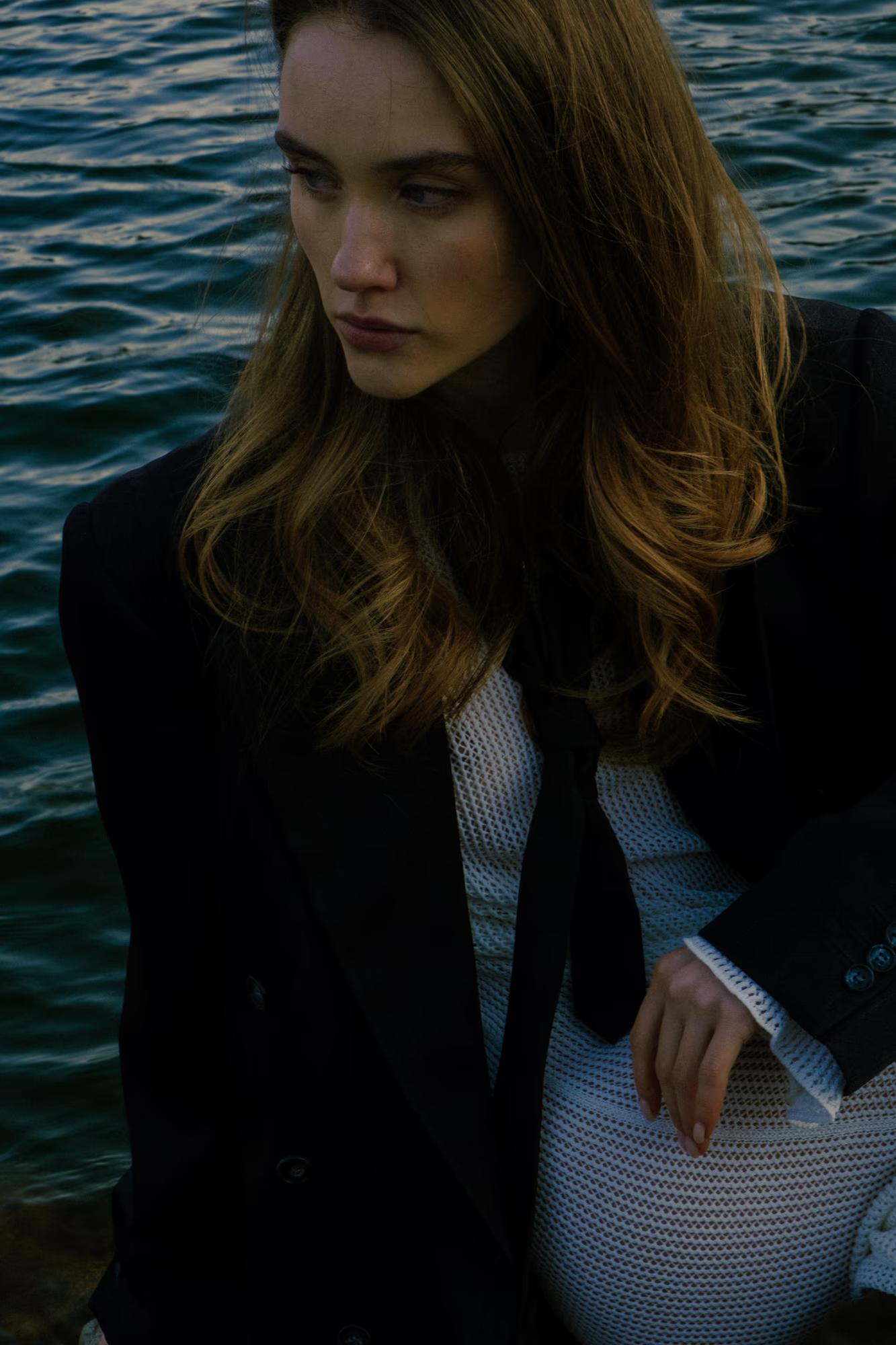 Medium shot: A female model in a black blazer and white top posing against a backdrop of a rippling lake at dusk.