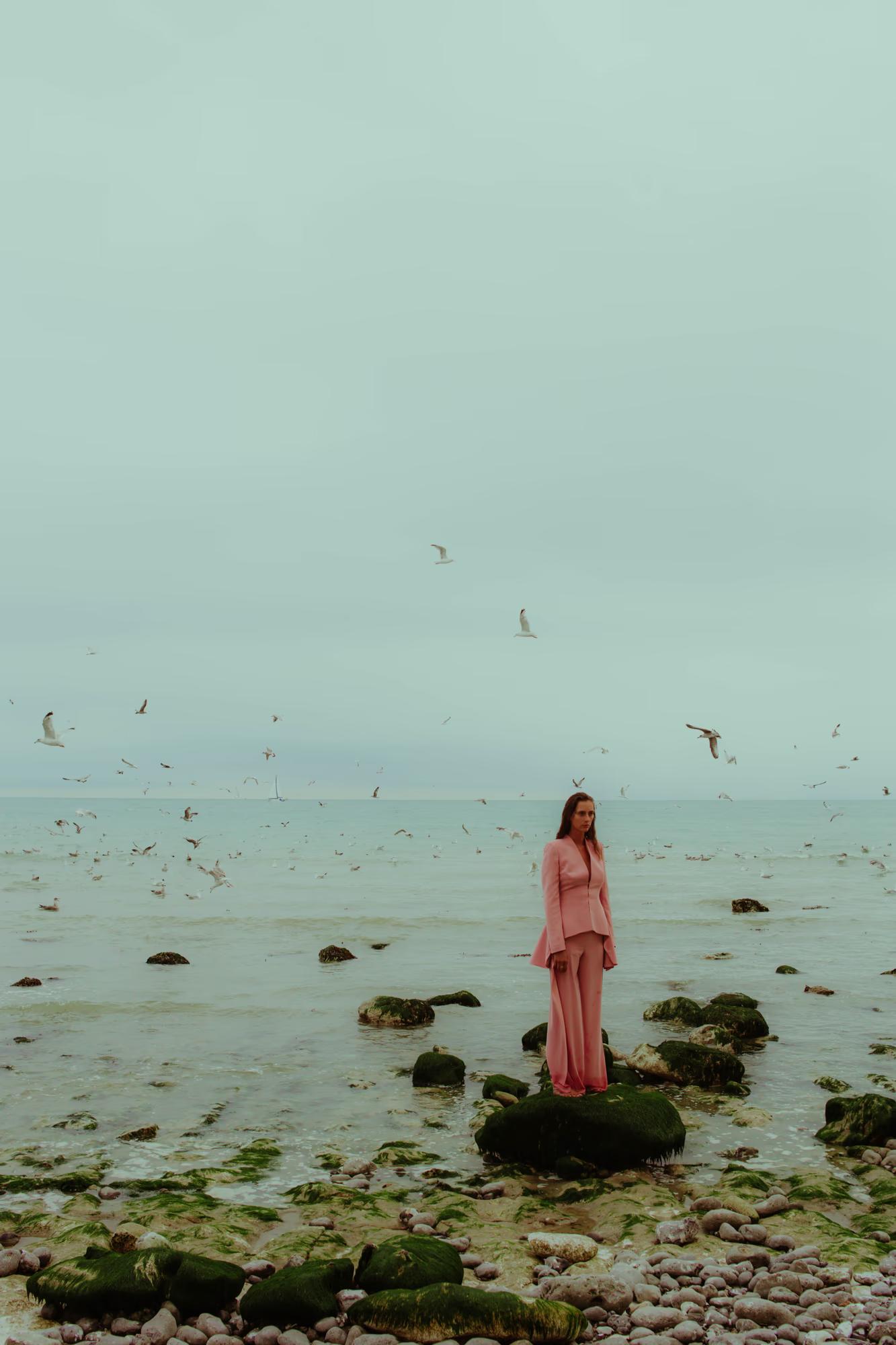 Full body fashion campaign shot of woman in red designer outfit standing on Eastbourne shoreline with calm sea and overcast sky, distant coastal editorial