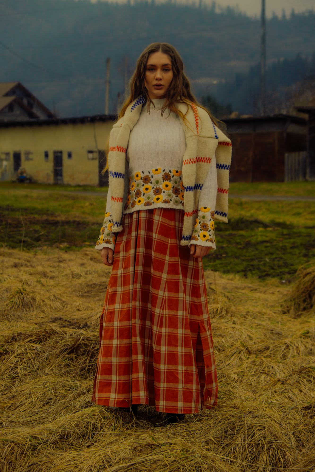 Full body fashion shot of woman in white blouse and bold red tartan maxi skirt in golden Carpathian meadow with mountain village visible, ethno style editorial