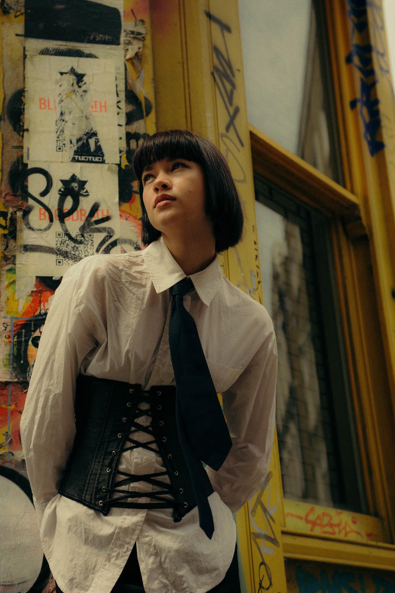Medium fashion shot of woman in white shirt with dark corset belt standing by window, androgynous tailored editorial, warm tones, Shoreditch London
