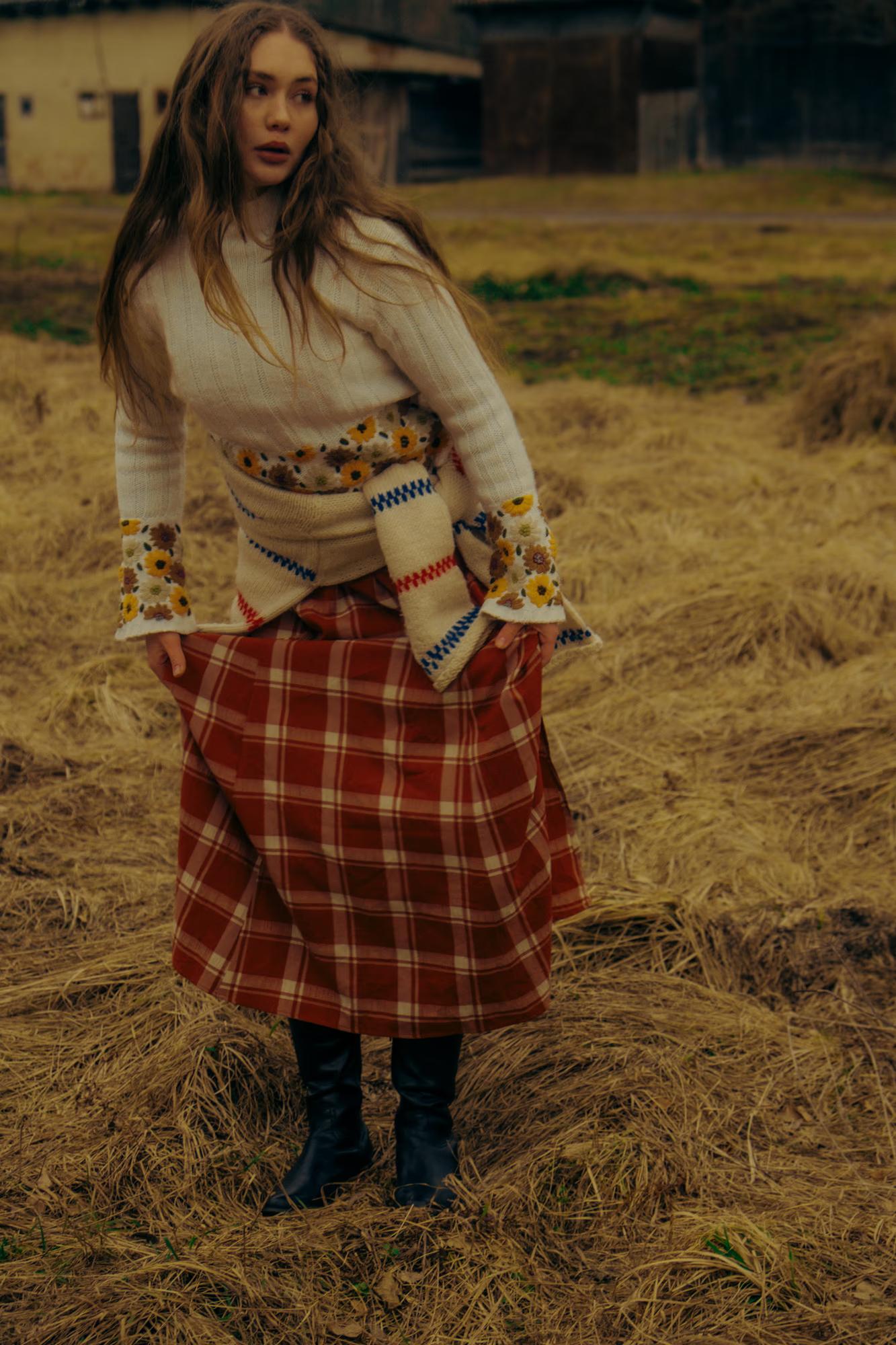 Full body fashion editorial of woman in white blouse and red plaid midi skirt standing in Carpathian mountain field, rustic ethno style, overcast natural light