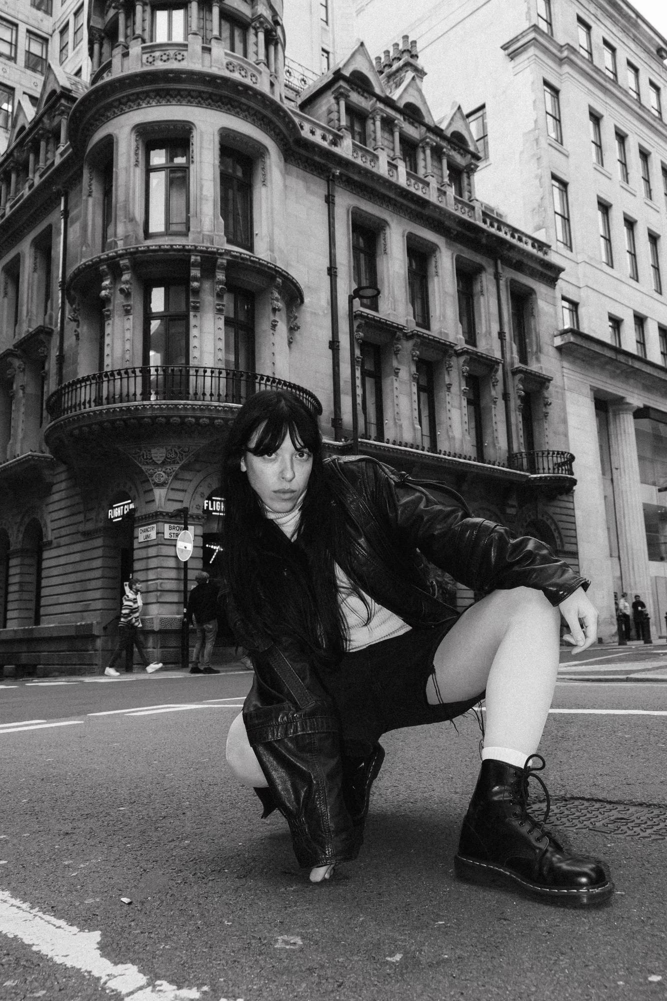 Full body fashion shot of person in dark layered outfit seated on Manchester street pavement with classic city architecture, warm tones, urban editorial