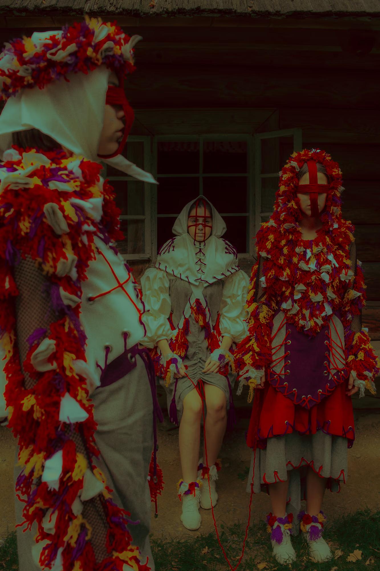 Group editorial shot of models in vibrant Ukrainian folk-inspired costumes with floral headpieces standing in front of a traditional wooden house.