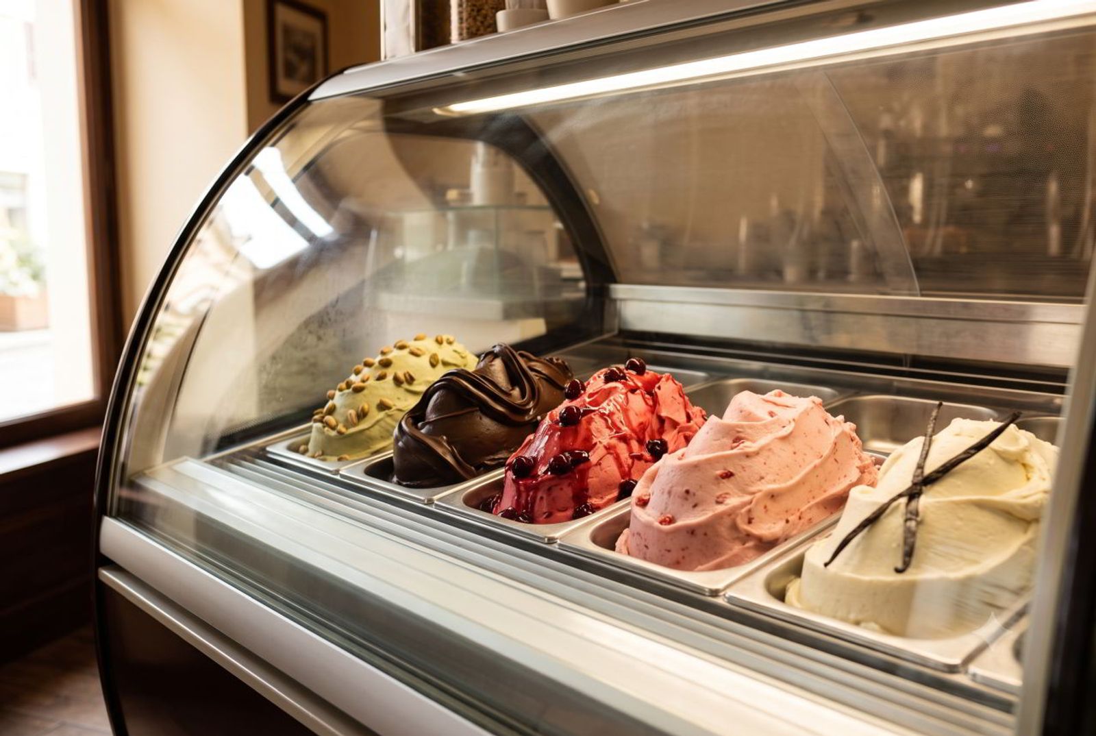 Decorated pans of pistachio, chocolate, amarena, strawberry, and vanilla gelato in a café display case.