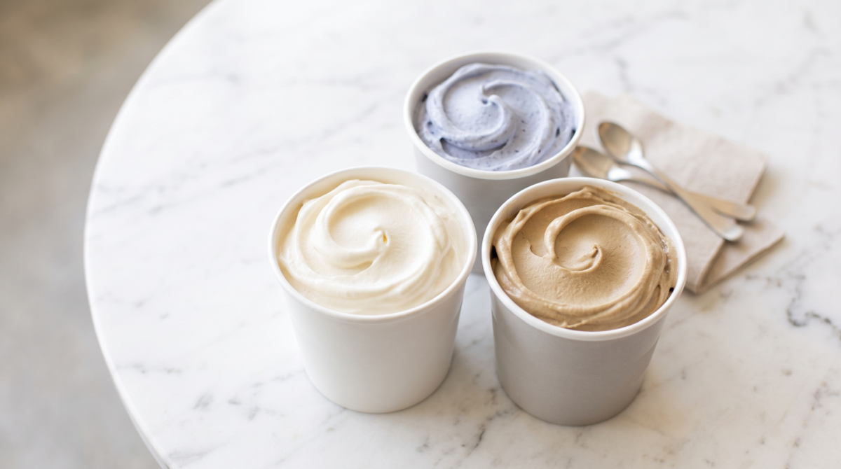 A close-up, top-down shot of three small, sleek tasting cups filled with distinctly different flavors of frozen yogurt—one creamy white (like vanilla or cheesecake), one pale blue (blueberry), and one light brown (coffee)