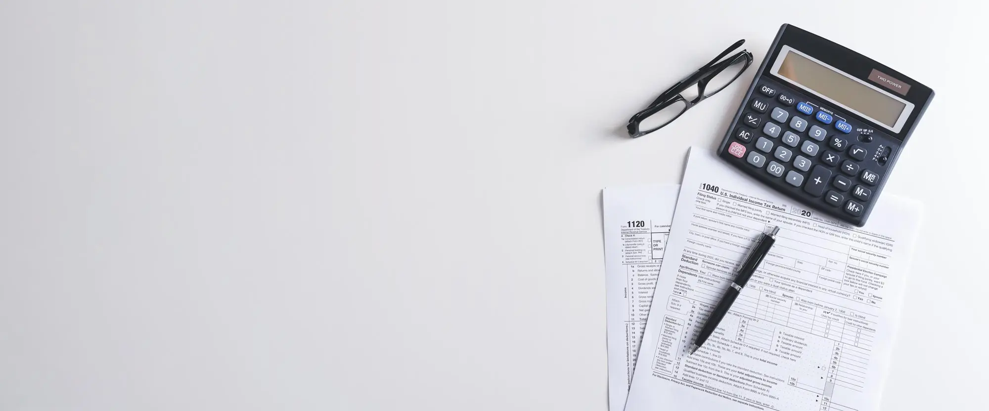 Blank IRS Form 1040 and Form 1120 tax returns on a white desk with a pen, reading glasses and a calculator.