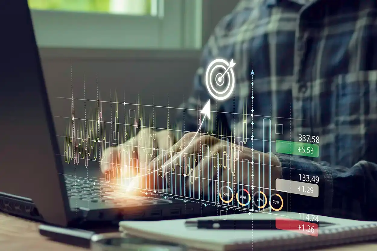Unas manos escribiendo en una computadora portátil con gráficas y cifras financieras superpuestas en la pantalla.