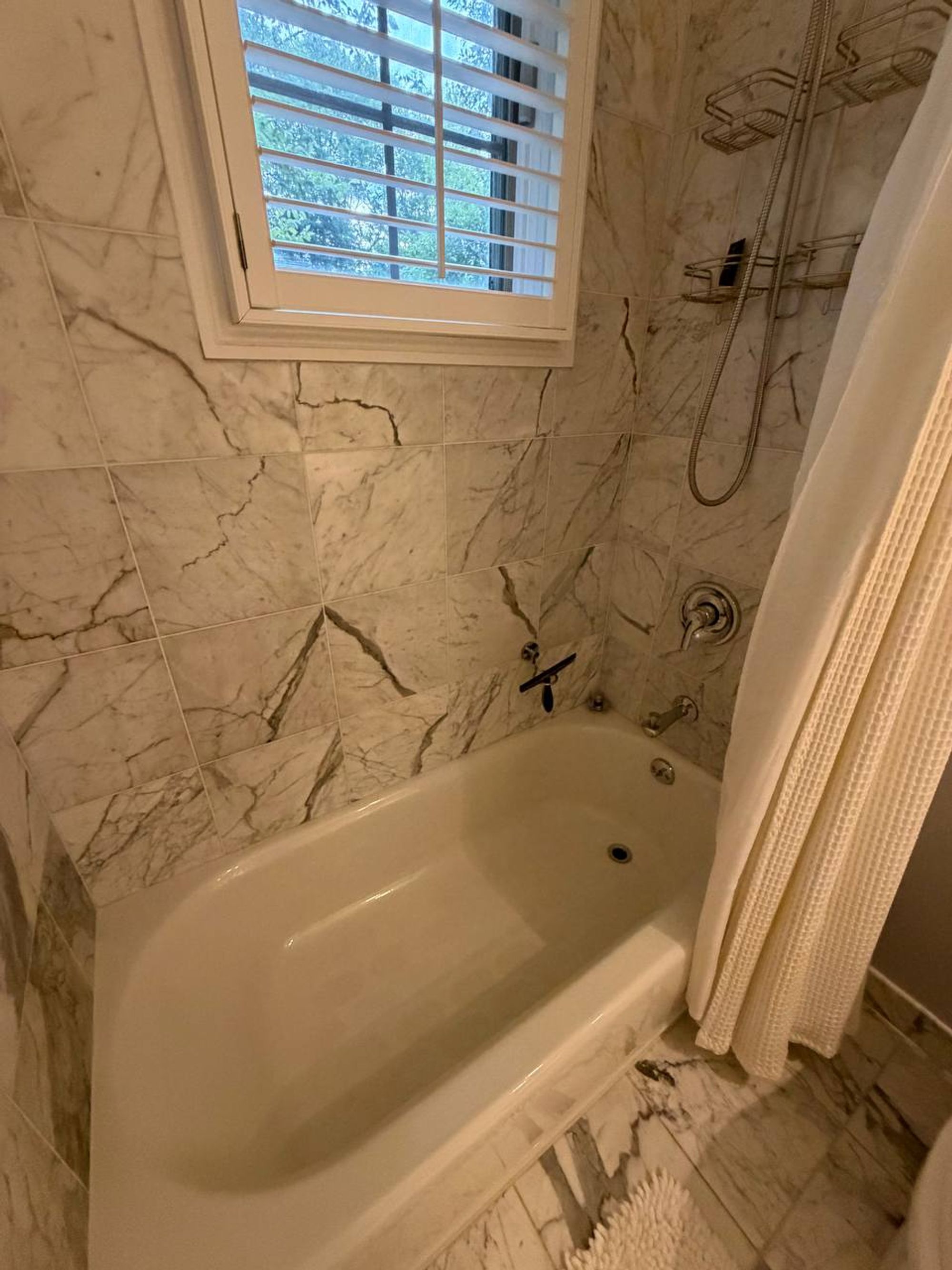 White soaking tub with gray veined marble walls and shuttered window