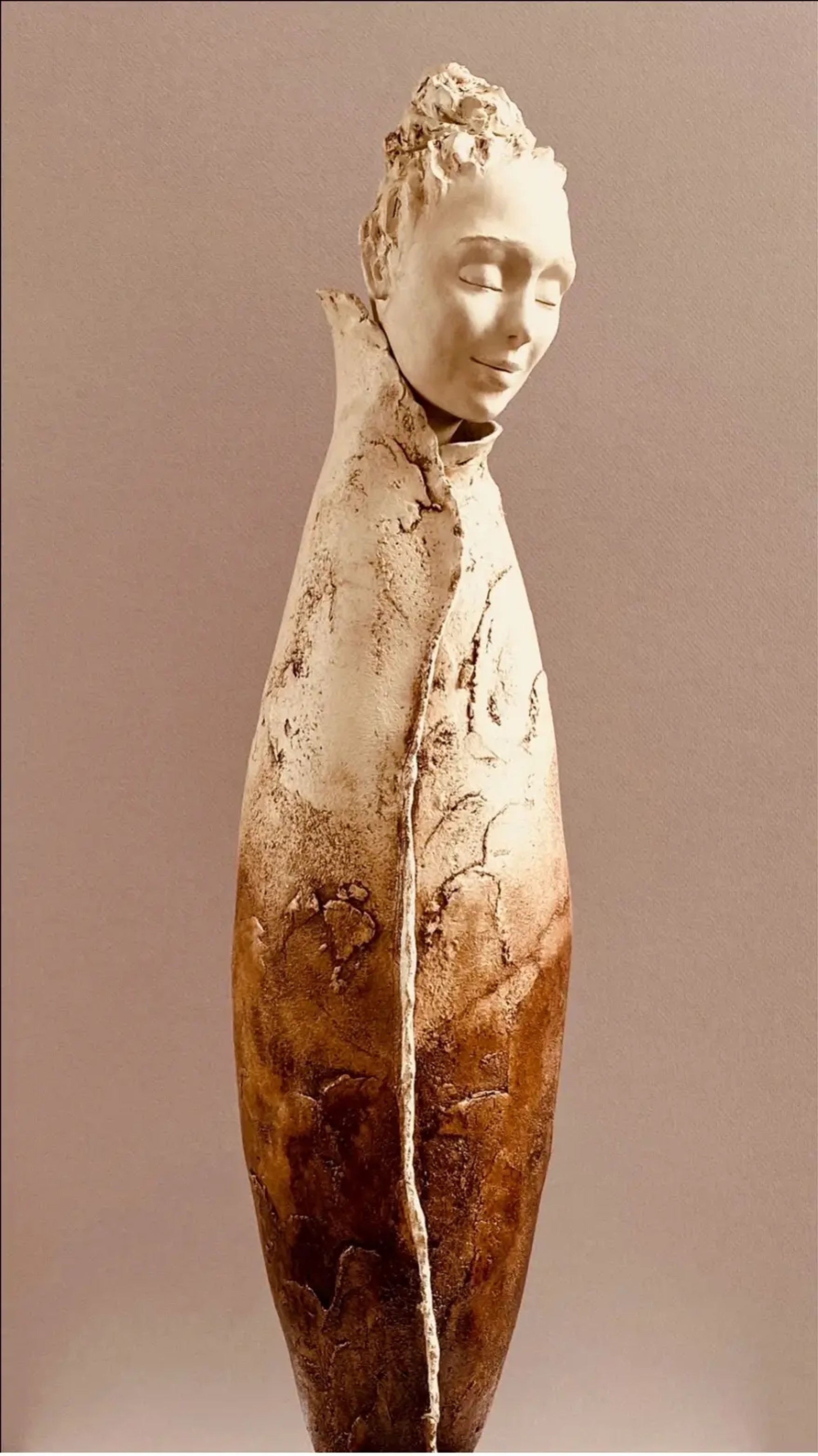 Sculpture élancée en grès, figure à visage serein émergeant d'une forme fuselée aux teintes dégradées d'ivoire et d'ocre, marquée de textures craquelées évoquant une métamorphose terrestre.
