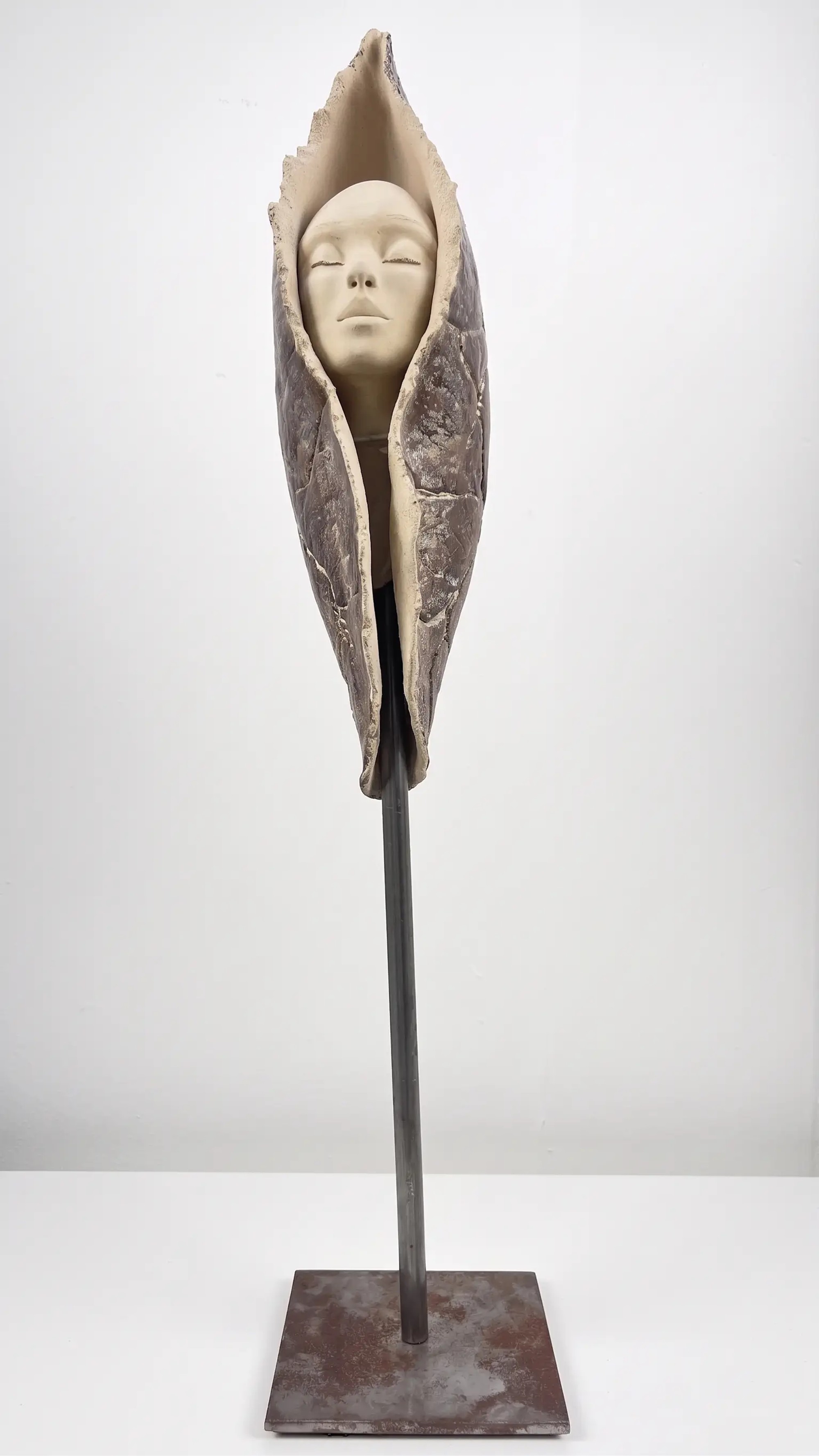 Sculpture en grès beige et brun sombre, visage apaisé enveloppé dans une forme organique verticale sur socle en métal noir.