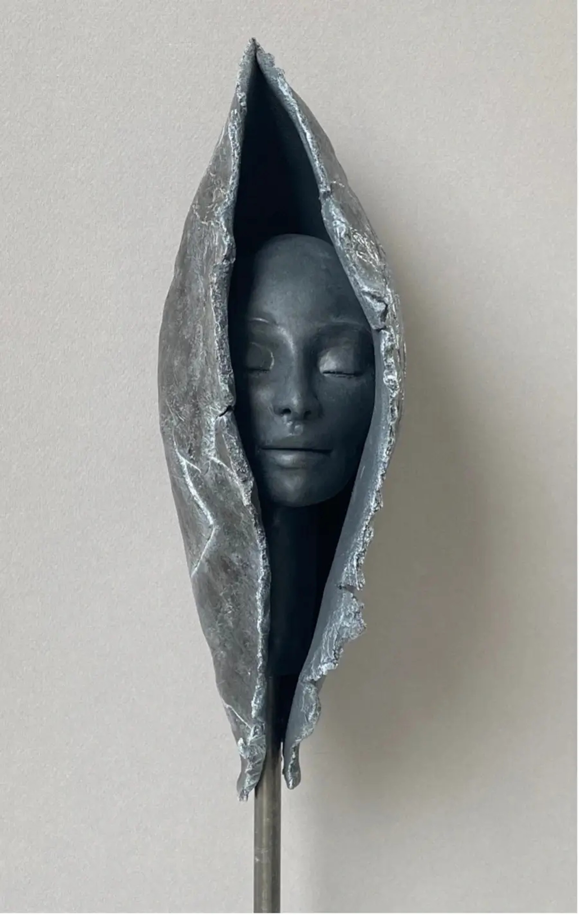 Sculpture en grès argenté, visage aux yeux clos émergeant d'une enveloppe texturée et brillante, évoquant l'éclosion d'une conscience depuis un cocon métallisé posé sur tige fine.