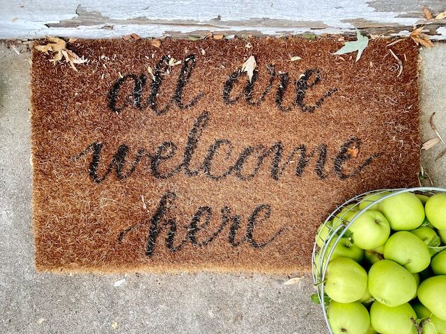 Door mat on a porch reading "all are welcome here"