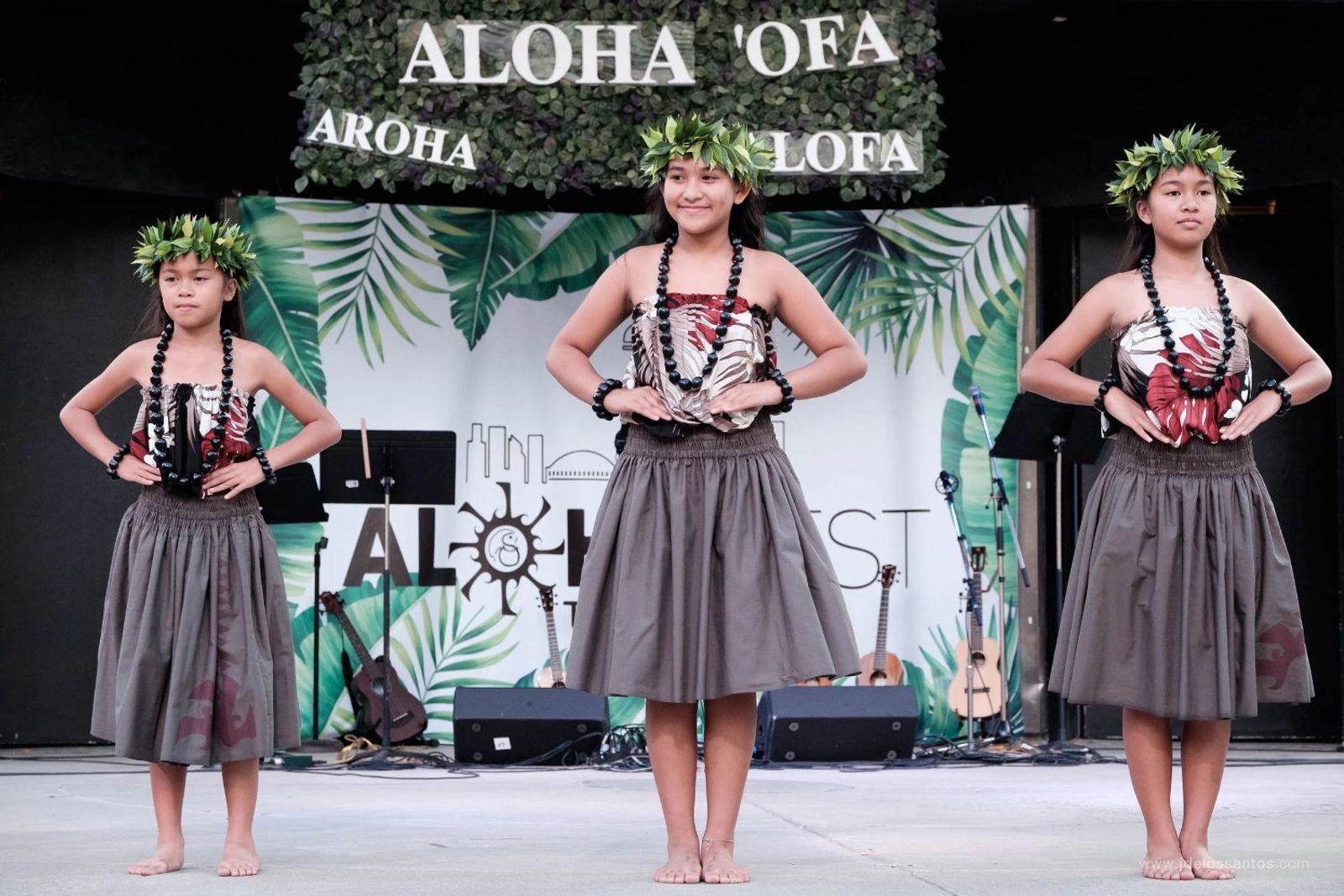 Three young hālau ʻōpio dancers in grey pāʻū and maile lei on the AlohaFest stage