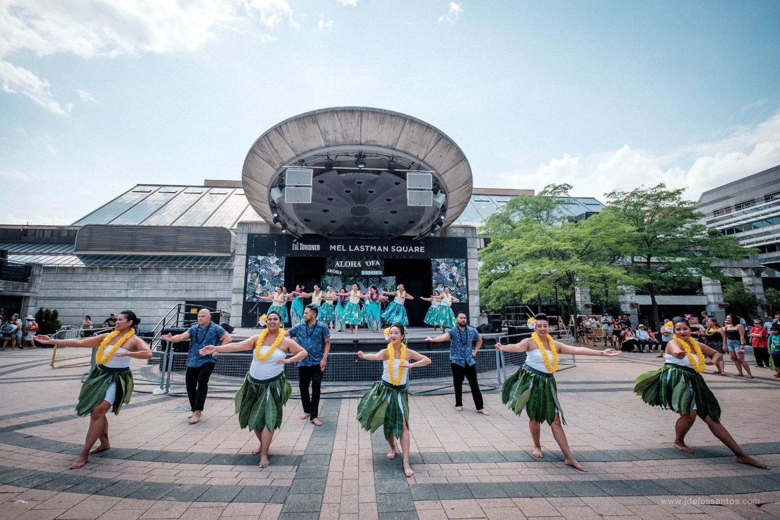 Hālau dancers in green ti leaf skirts performing kahiko with arms extended