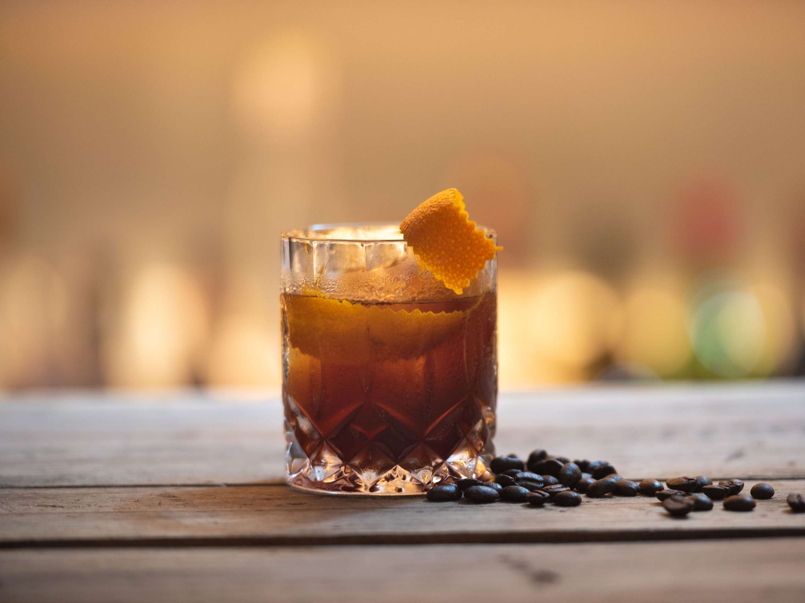 Illustrative photograph of a dark amber drink over ice in a cut-glass tumbler on a wooden bar