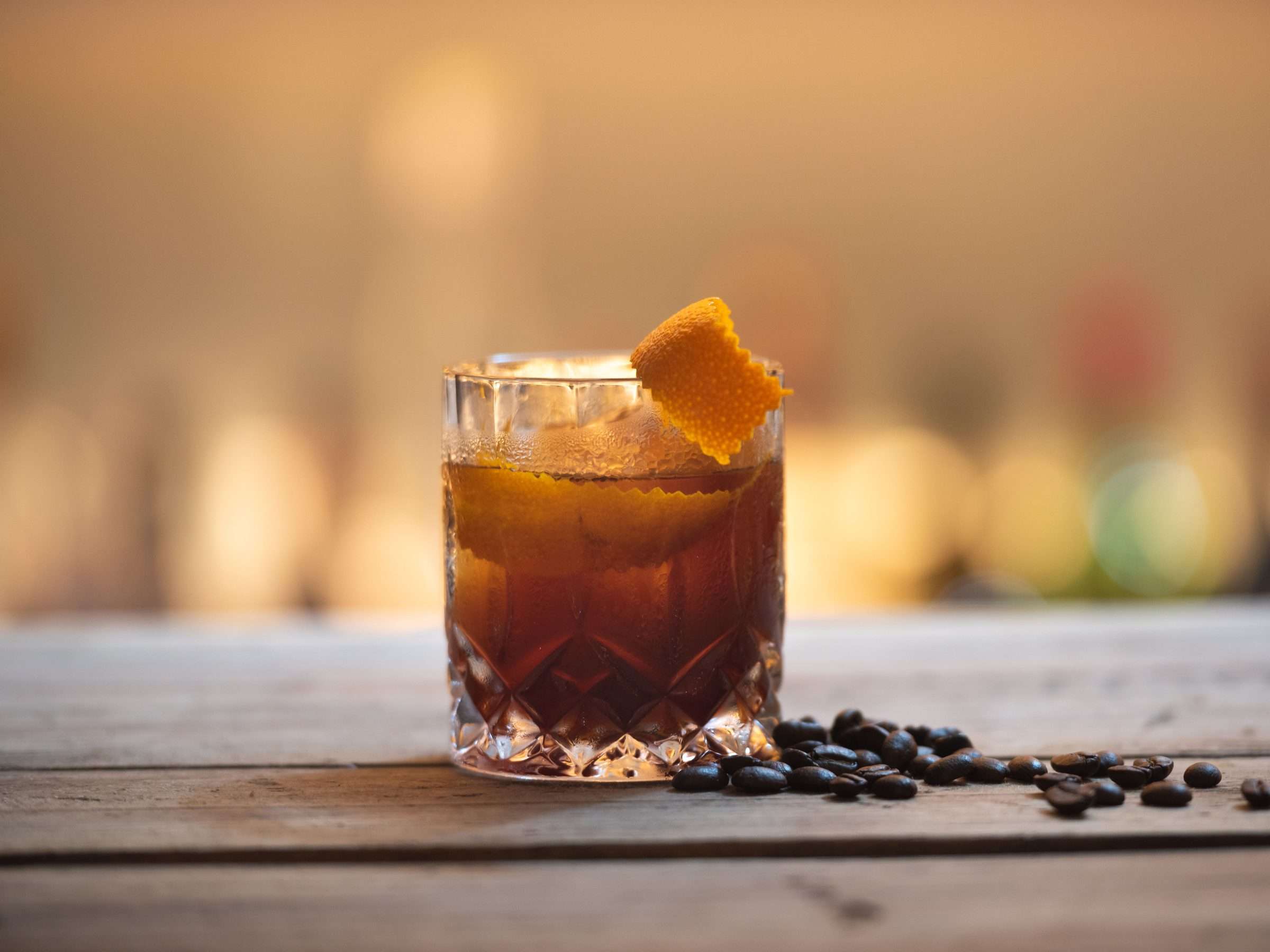 Illustrative photograph of a dark amber drink over ice in a cut-glass tumbler on a wooden bar