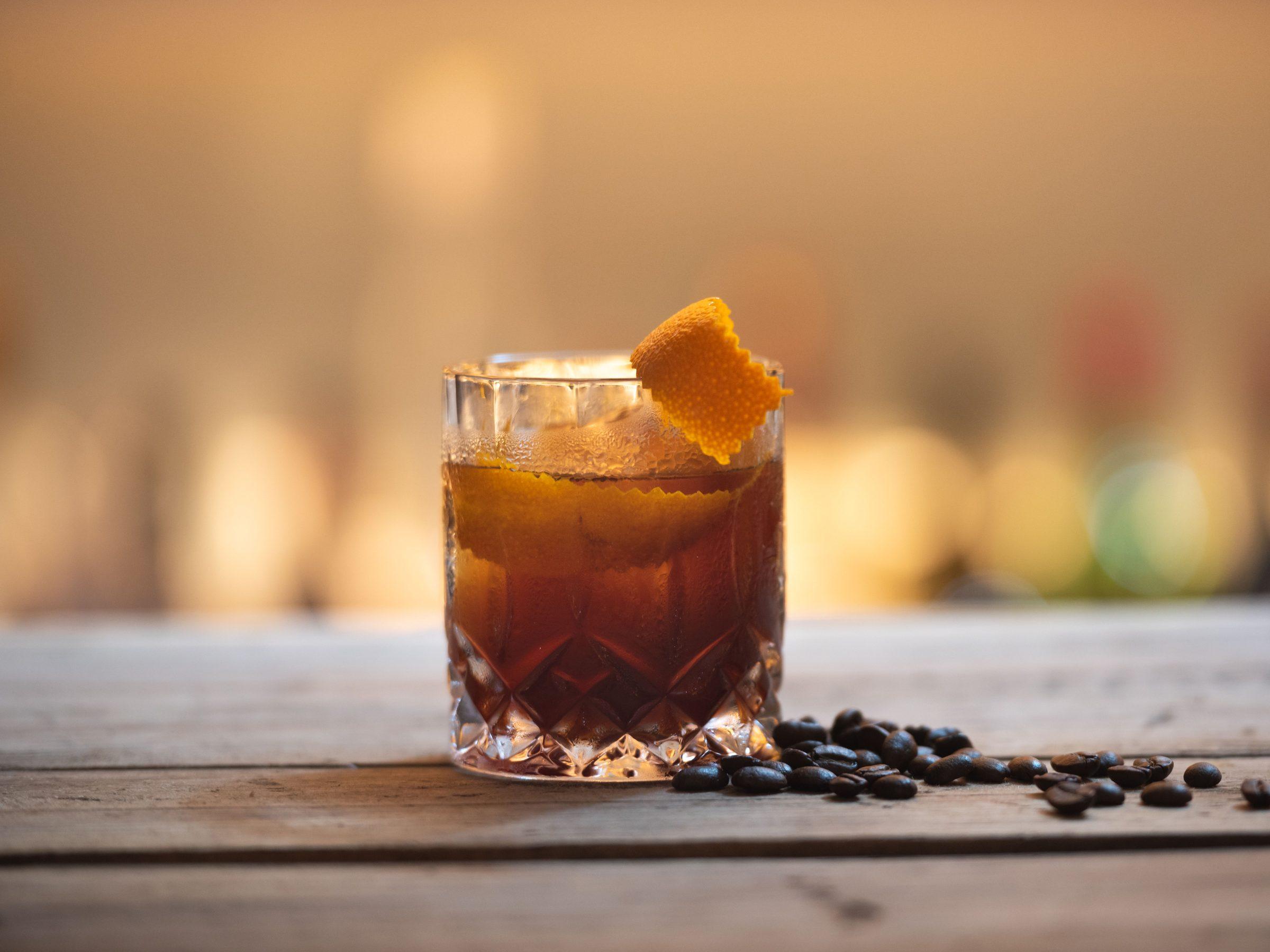 Illustrative photograph of a dark amber drink over ice in a cut-glass tumbler on a wooden bar
