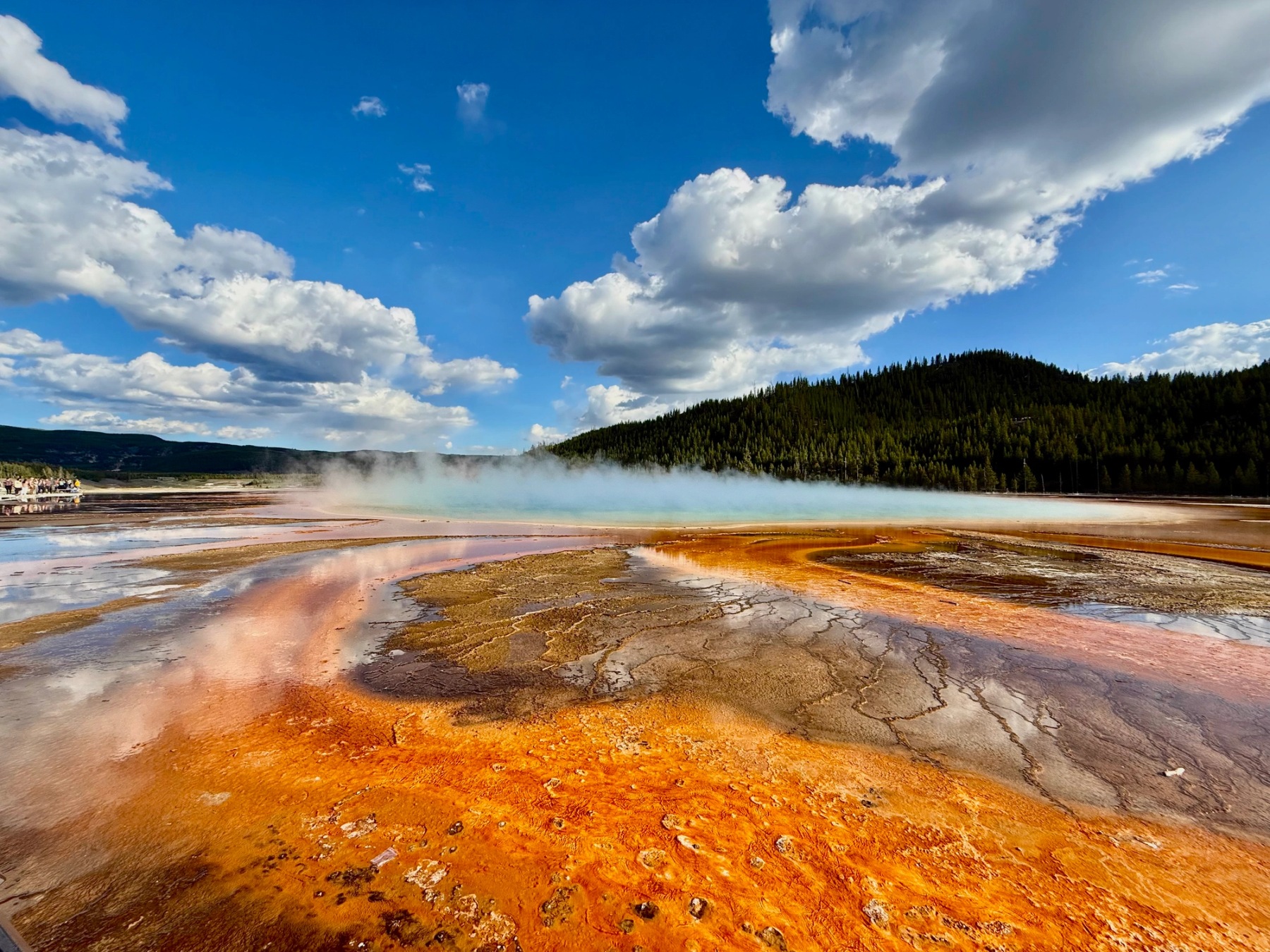 Orange geothermal terrain and steaming blue water beneath dramatic clouds and forested hills in Yellowstone National Park.