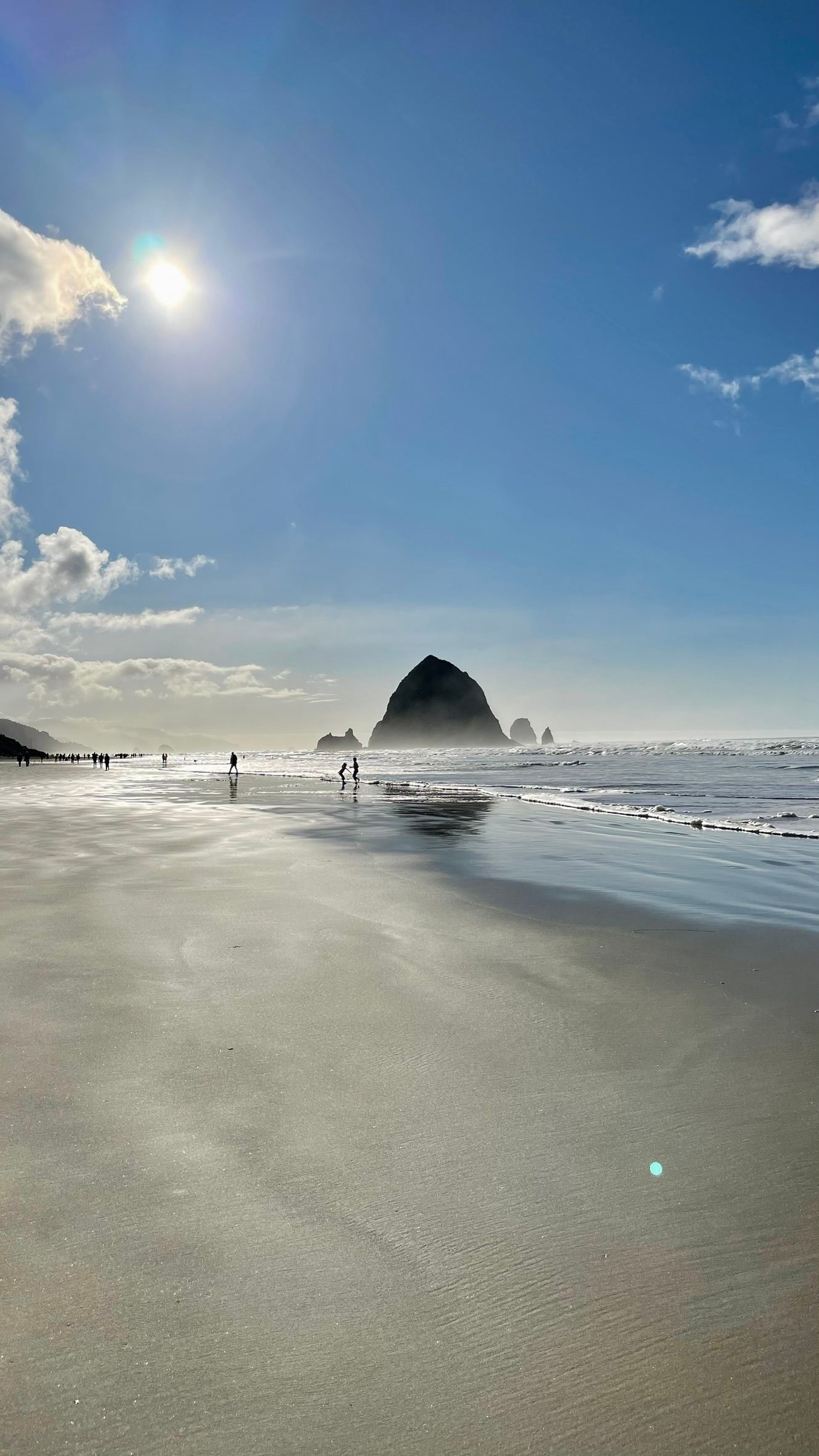 Haystack Rock at Cannon Beach reflected across wet sand beneath a bright blue sky, with the sun above the shoreline and small figures walking near the water.