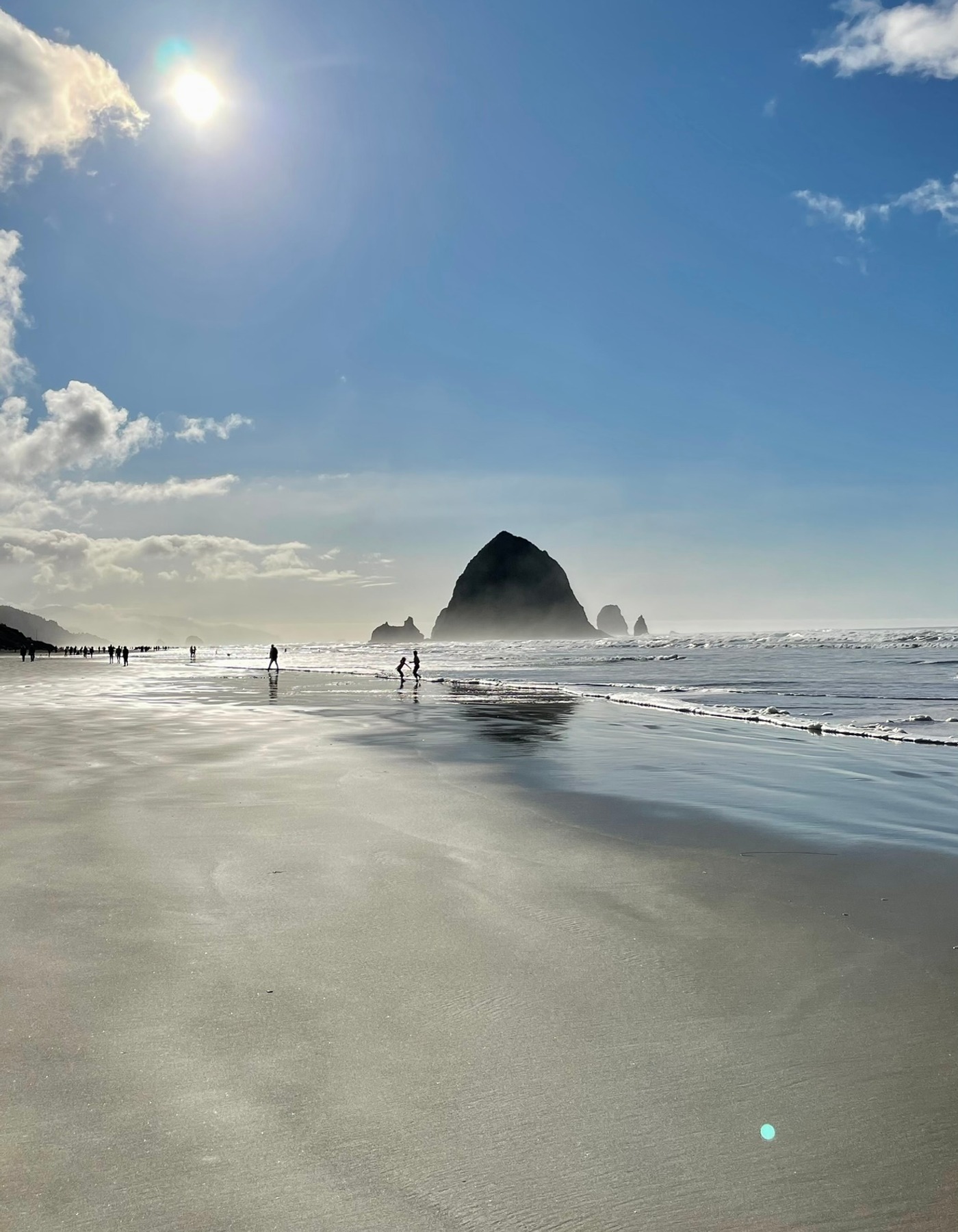 Haystack Rock at Cannon Beach reflected across wet sand beneath a bright blue sky, with the sun above the shoreline and small figures walking near the water.