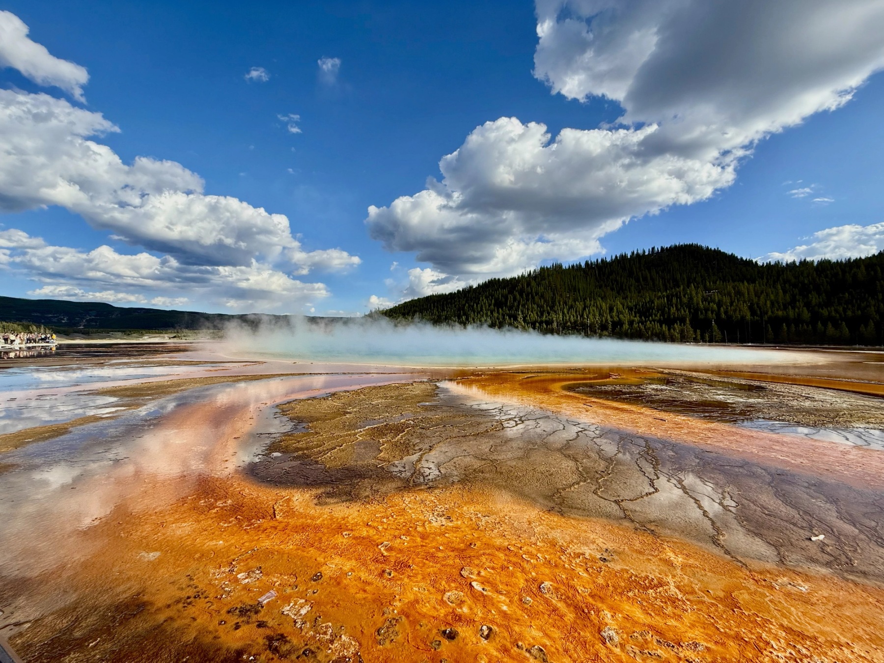 Orange geothermal terrain and steaming blue water beneath dramatic clouds and forested hills in Yellowstone National Park.