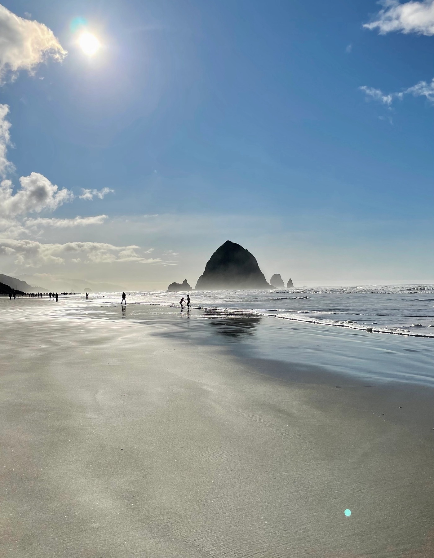 Haystack Rock at Cannon Beach reflected across wet sand beneath a bright blue sky, with the sun above the shoreline and small figures walking near the water.