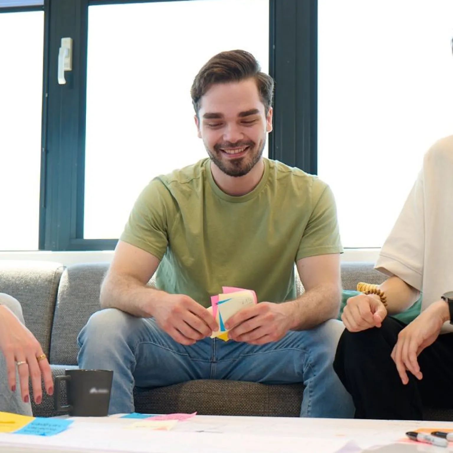 Man in een groen T-shirt glimlacht terwijl hij post-its vasthoudt, zittend op een bank.