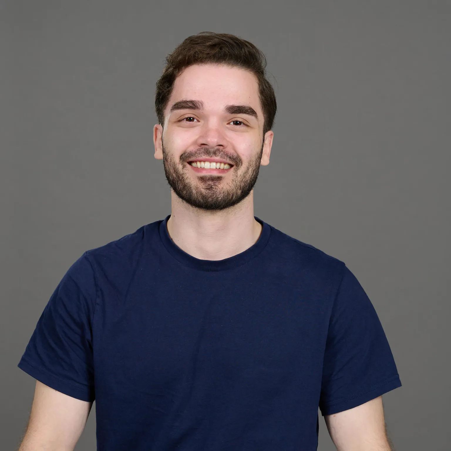 Glimlachende man met bruin haar en baard in een donkerblauw T-shirt tegen een grijze achtergrond.