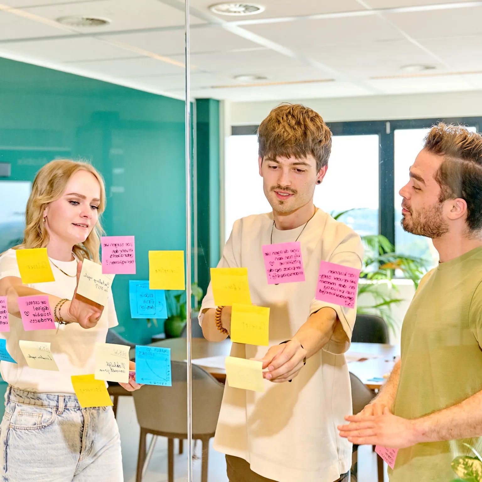 Drie mensen brainstormen samen bij een glazen wand met gekleurde post-its.