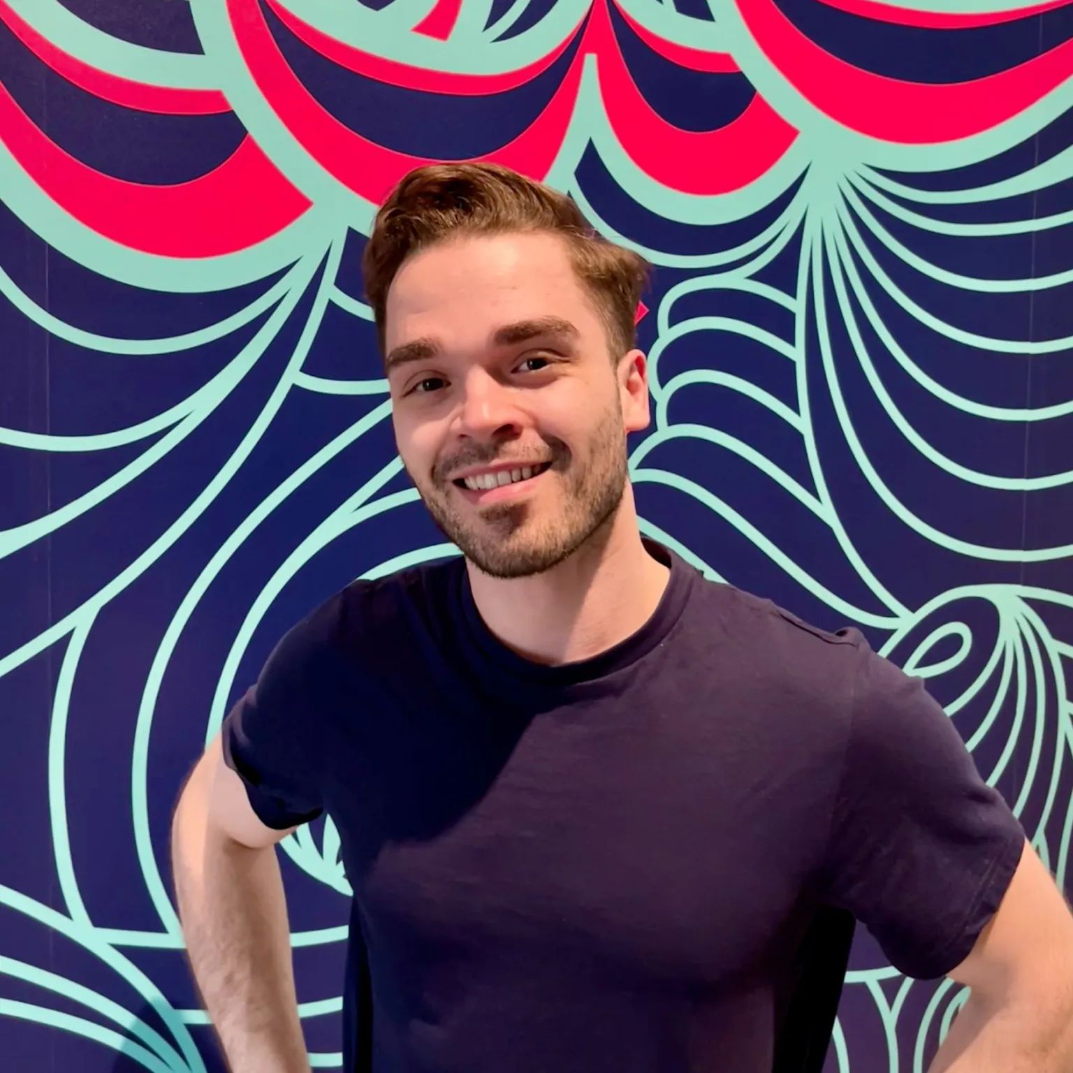Man in een donkerblauw T-shirt glimlacht voor een kleurrijke achtergrond met abstracte patronen.