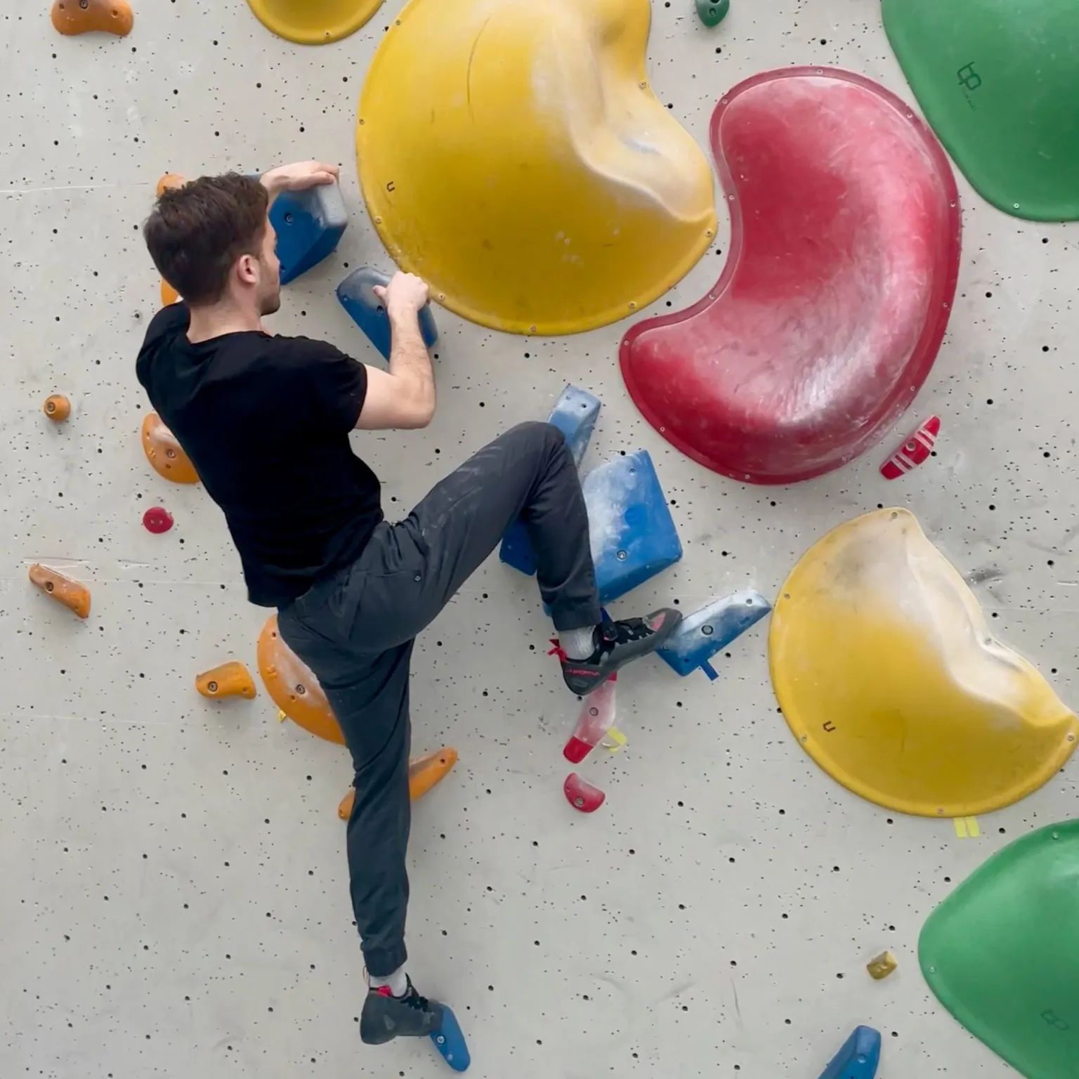Man in een zwart T-shirt klimt op een indoor klimmuur met kleurrijke grepen.