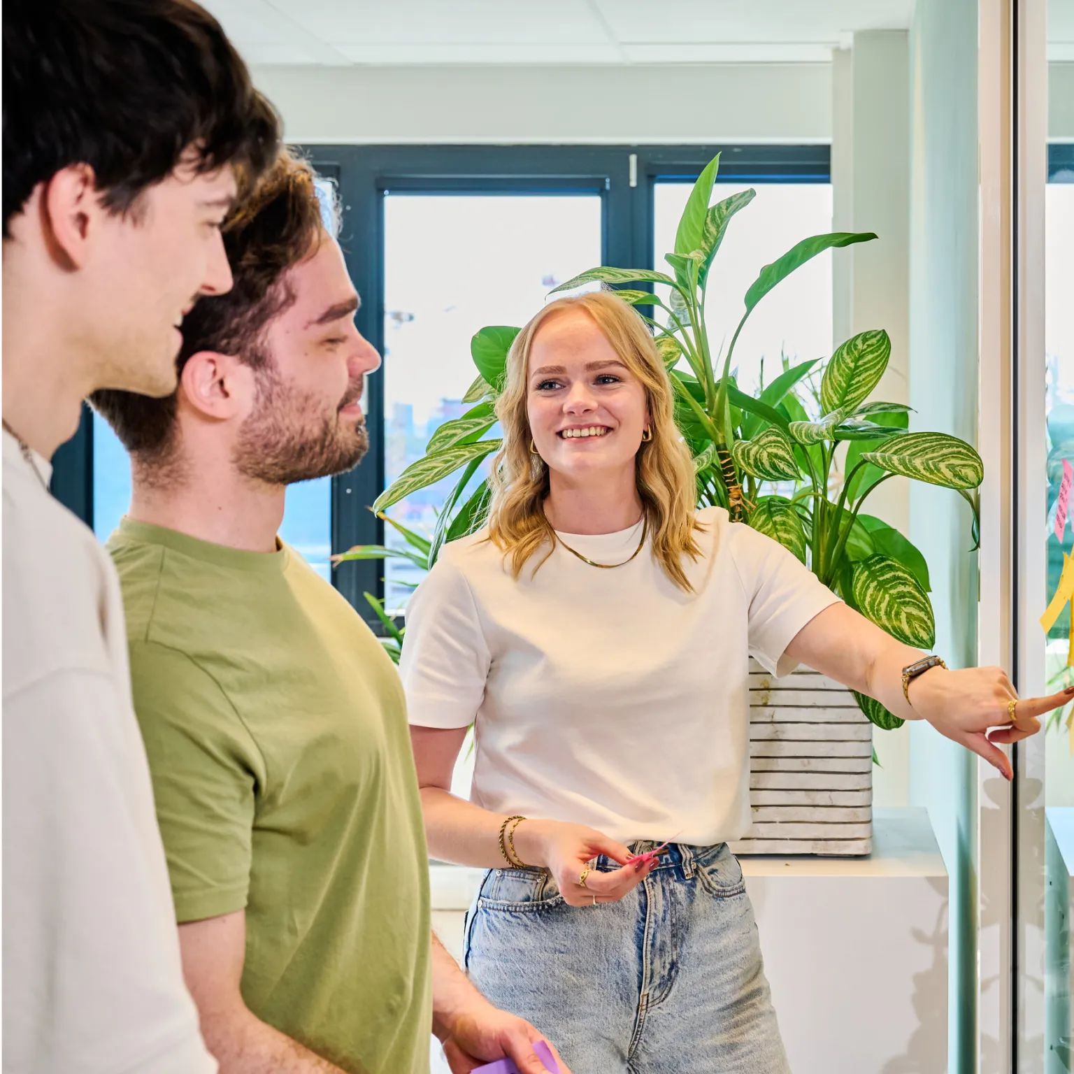Groep mensen overlegt terwijl een vrouw in een witte trui glimlachend naar een glazen wand wijst.