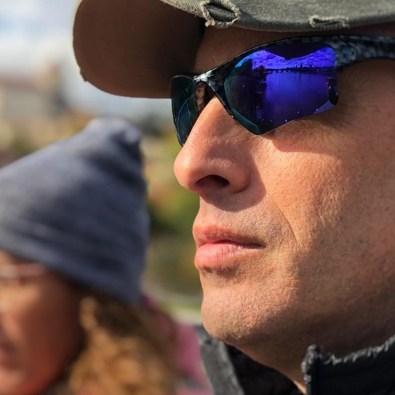 Side view of a man in a cap and reflective sunglasses, reflecting a bridge over water, with another person blurred in the background.