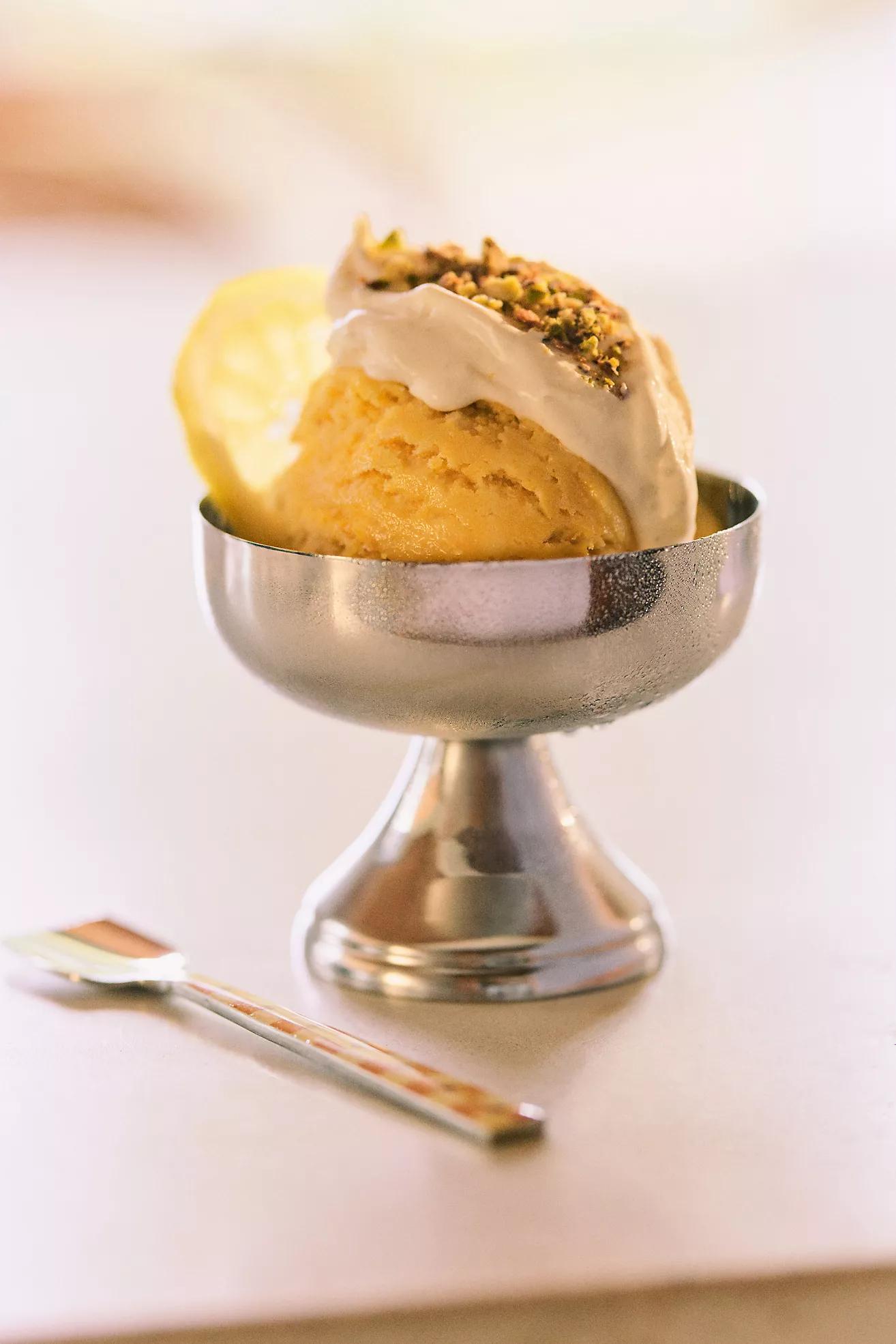 A metal, footed, dessert dish with a scoop of vanilla ice cream with a pistachio topping. 